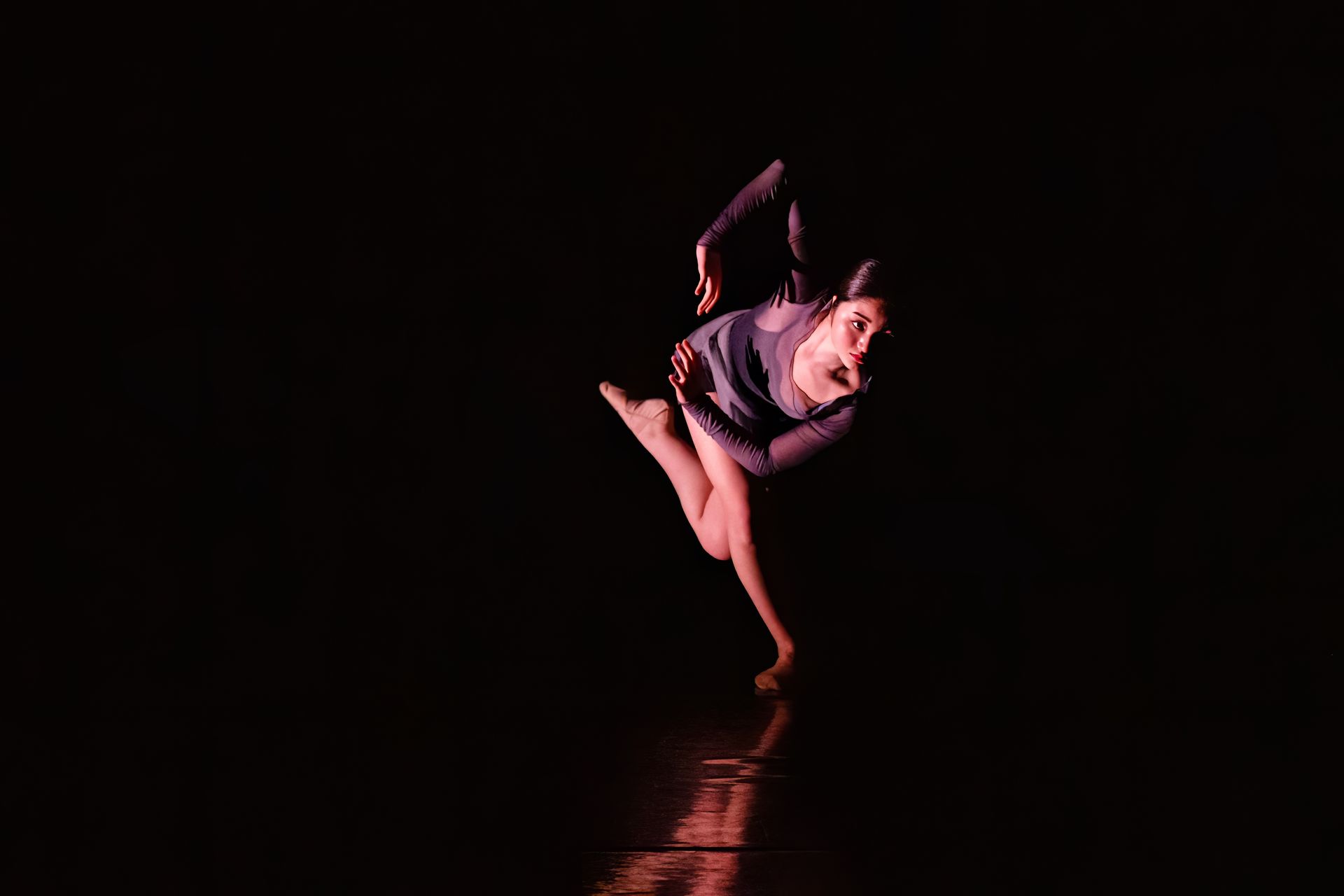 Dancer in purple costume striking a pose against a black background, lit by warm spotlight.
