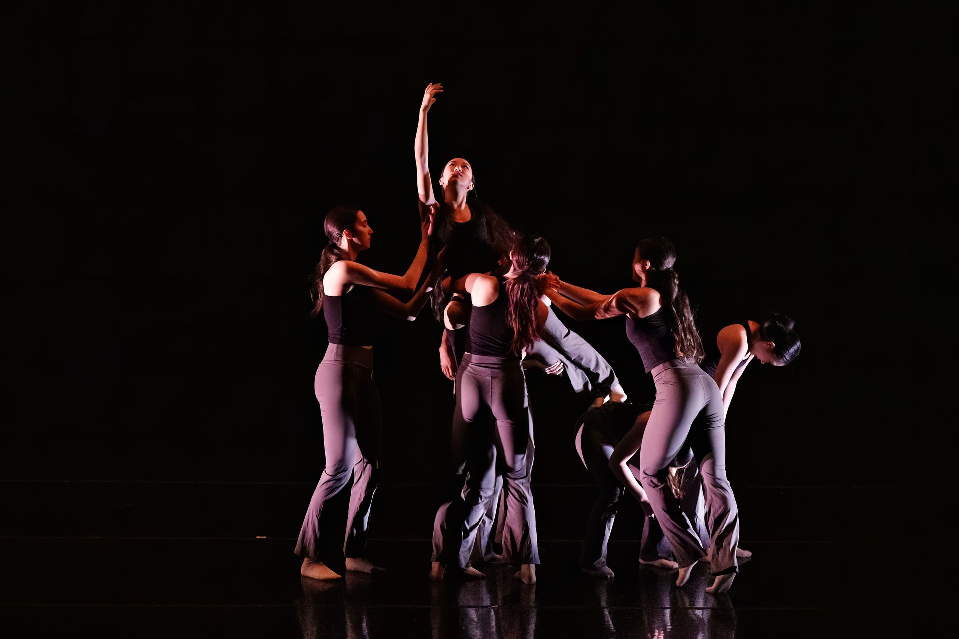 Dancers in dark costumes pose and move in a dramatic group formation on a black stage, lit with warm highlights