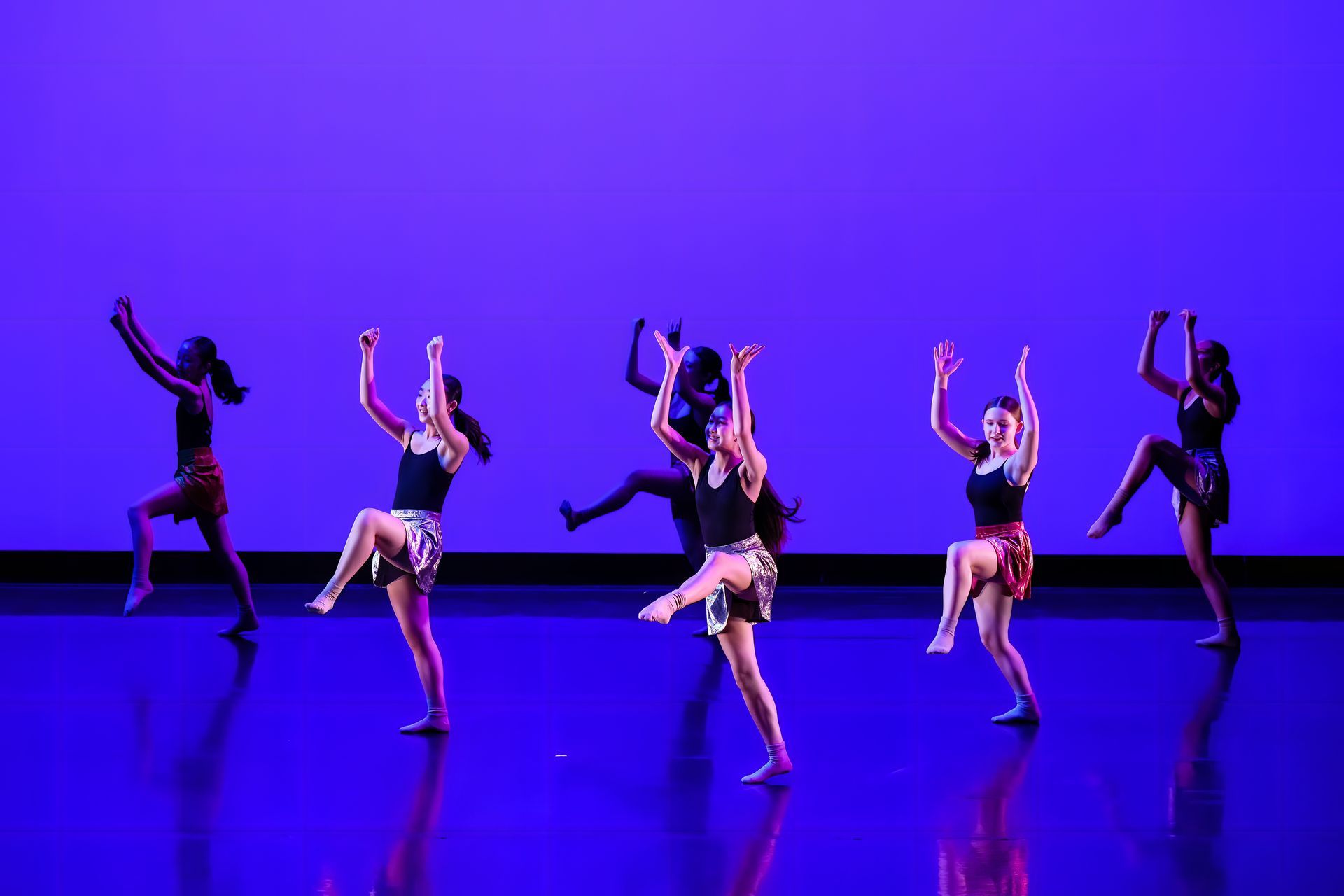 Five dancers in black and silver costumes perform on a purple-lit stage.
