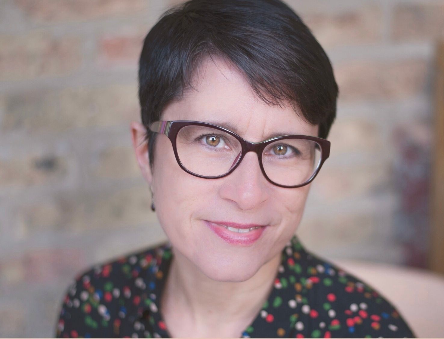 Close-up portrait of a person with short dark hair and glasses, smiling in front of a brick wall.