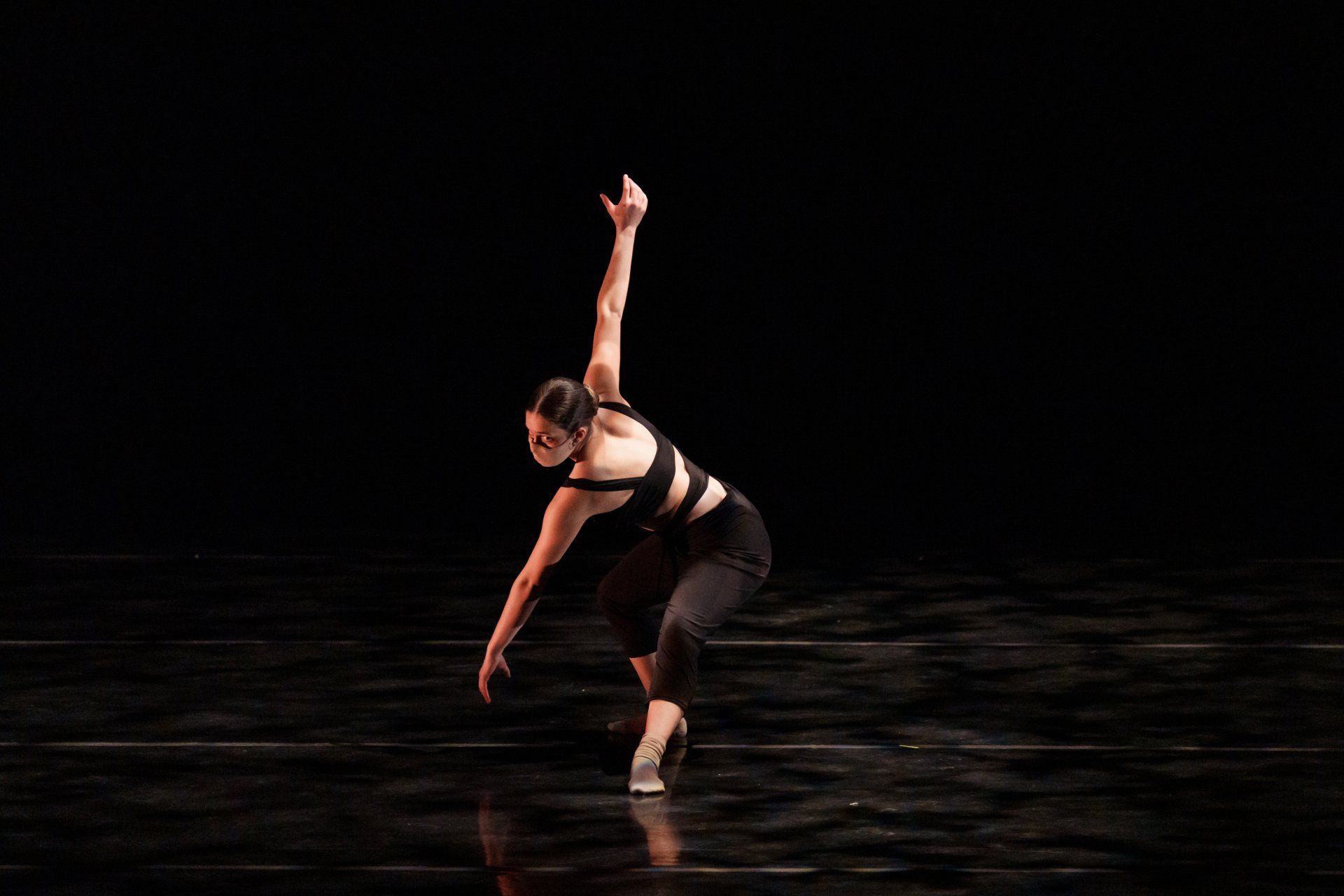 Dancer in a black costume striking a dramatic pose on a dark stage with reflective floor