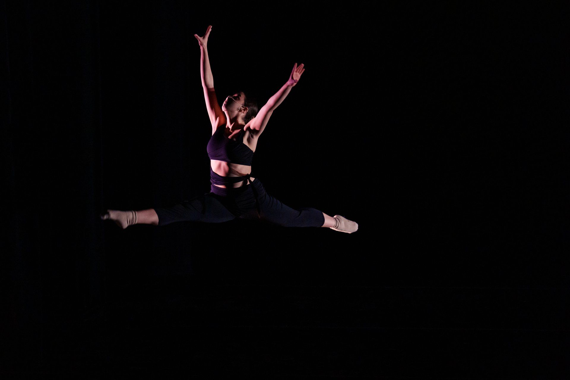 Dancer in a black outfit performing a split leap against a dark background.