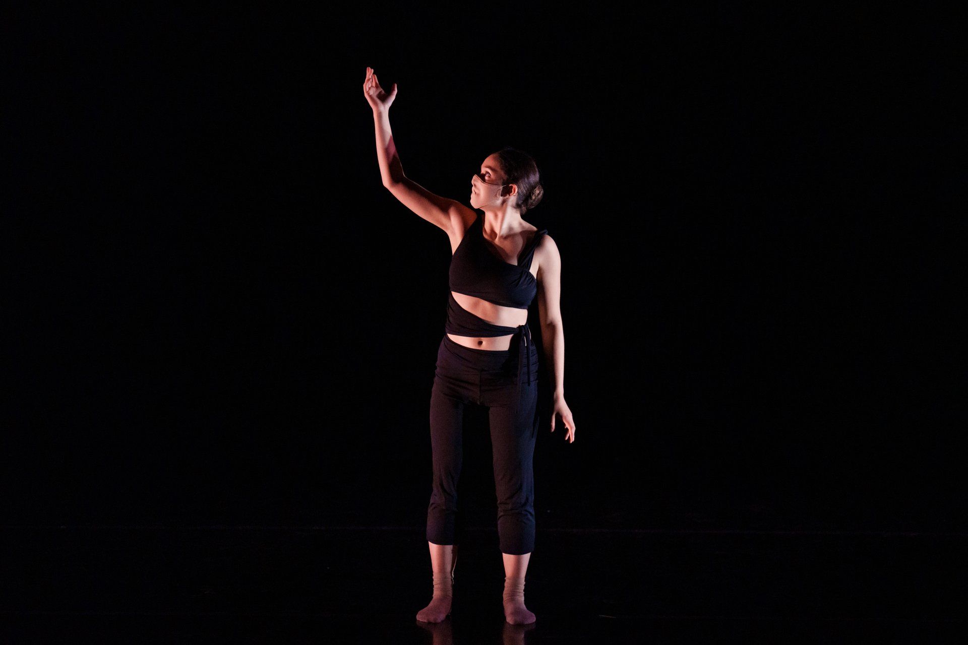 Person standing on a dark stage with one arm raised, wearing black clothing and a red top.