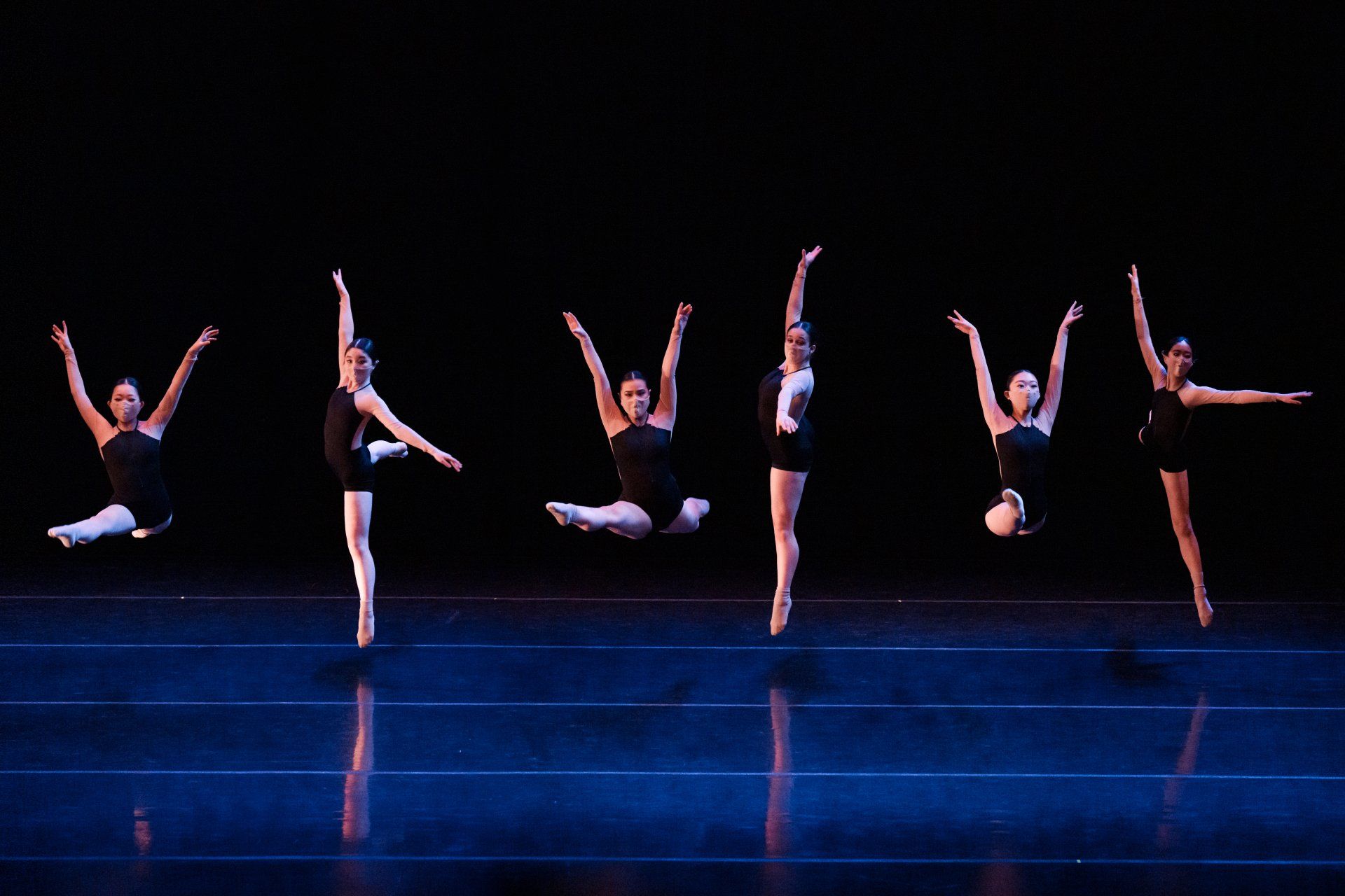 Six dancers in black costumes striking ballet poses on a dark blue stage