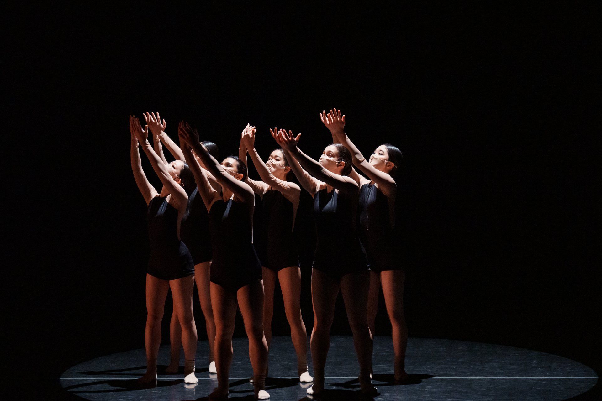 Five dancers in black outfits pose with arms raised on a dark stage under a spotlight.