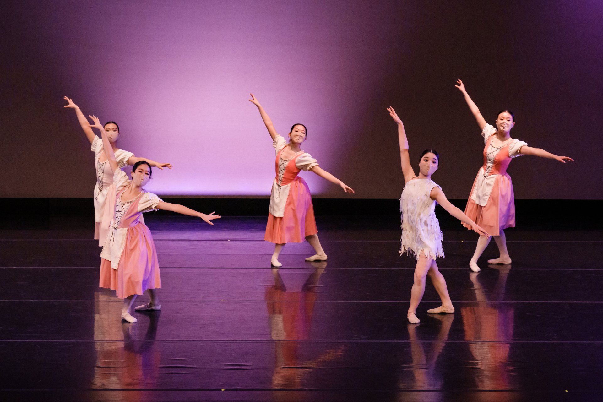 Six dancers in pink costumes pose on a stage with arms raised under purple lighting.