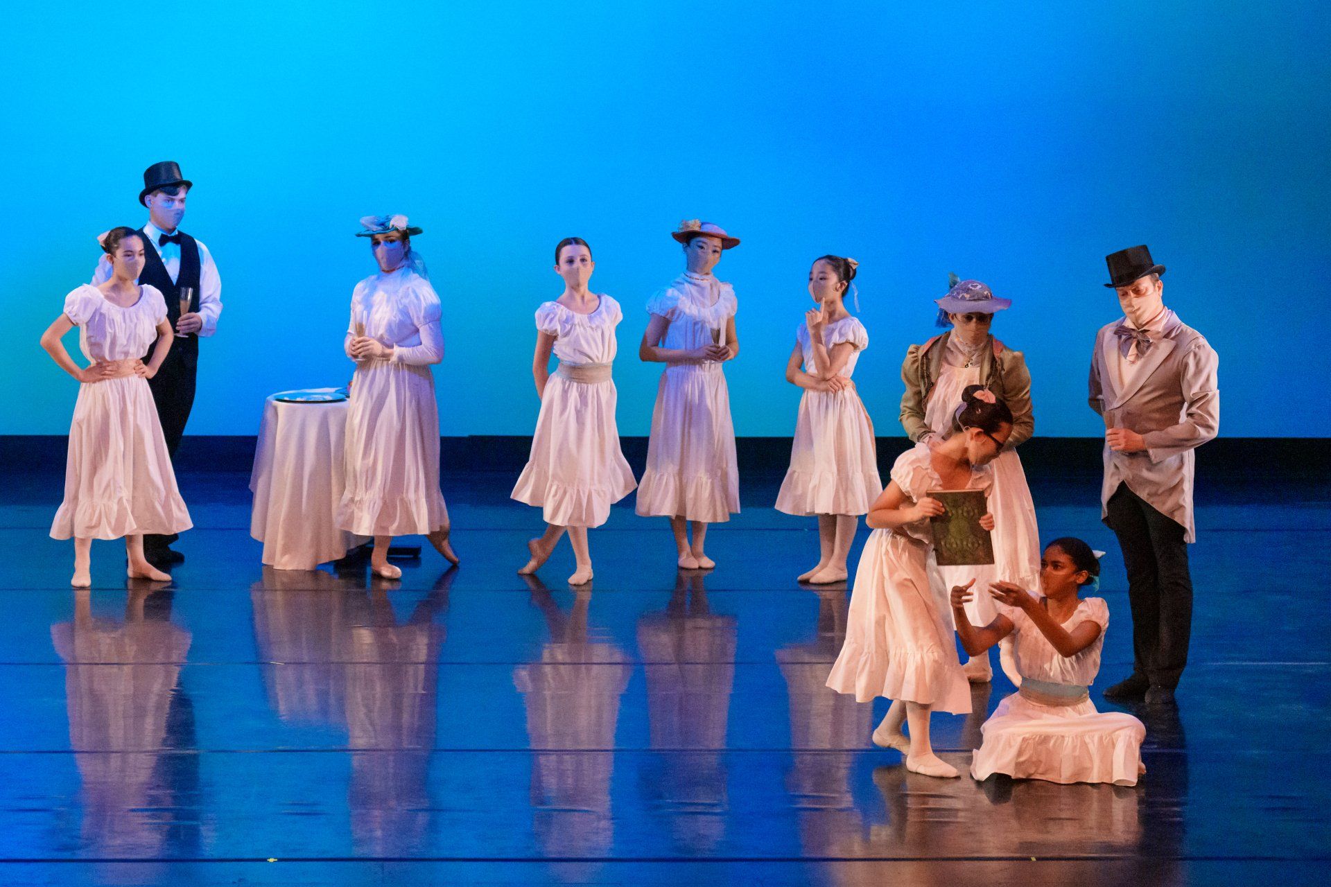 Dancers in white costumes on a blue-lit stage, with one seated pair in the foreground.