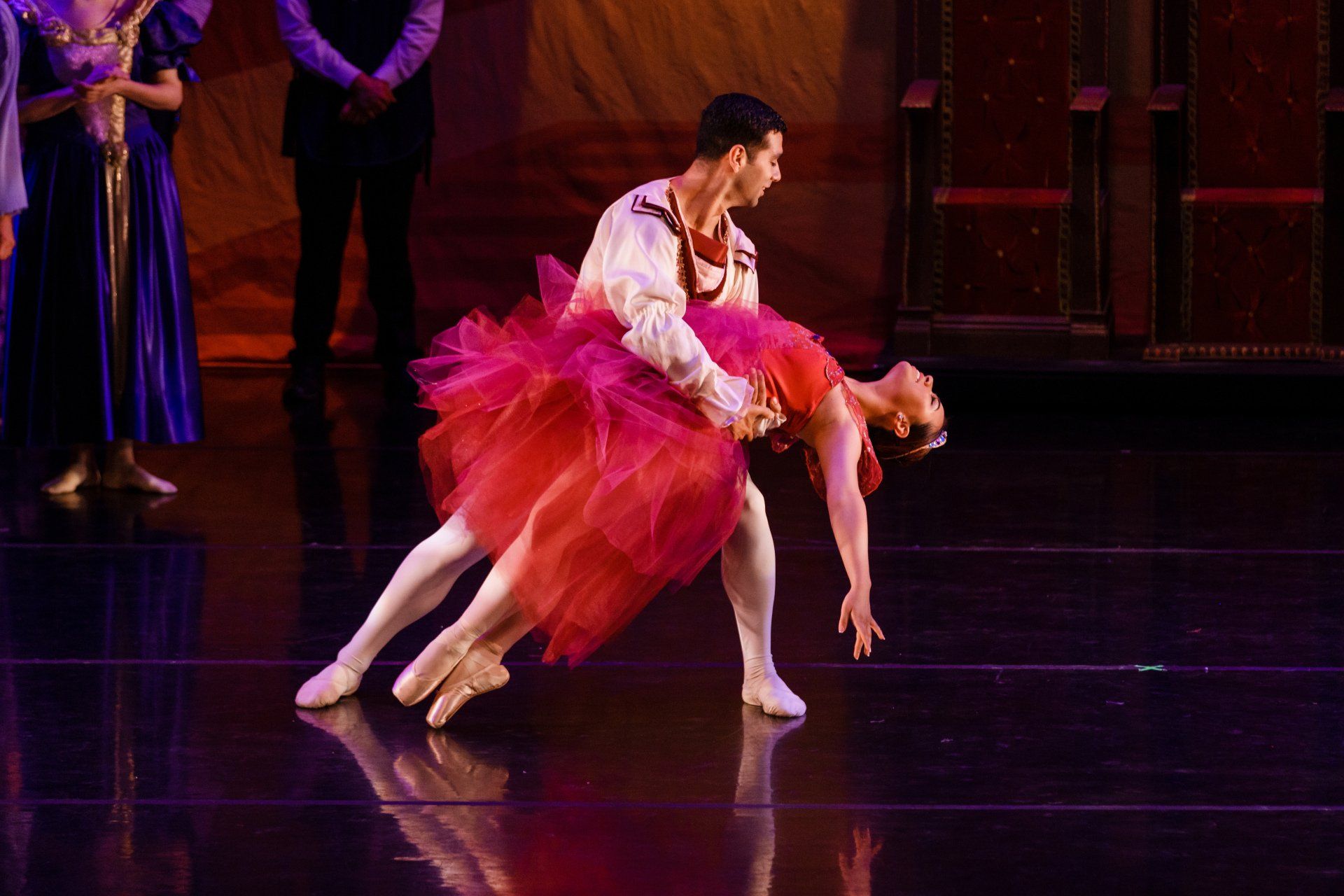 Two ballet dancers in a pink tutu perform a dramatic lift on a stage with a dark background.