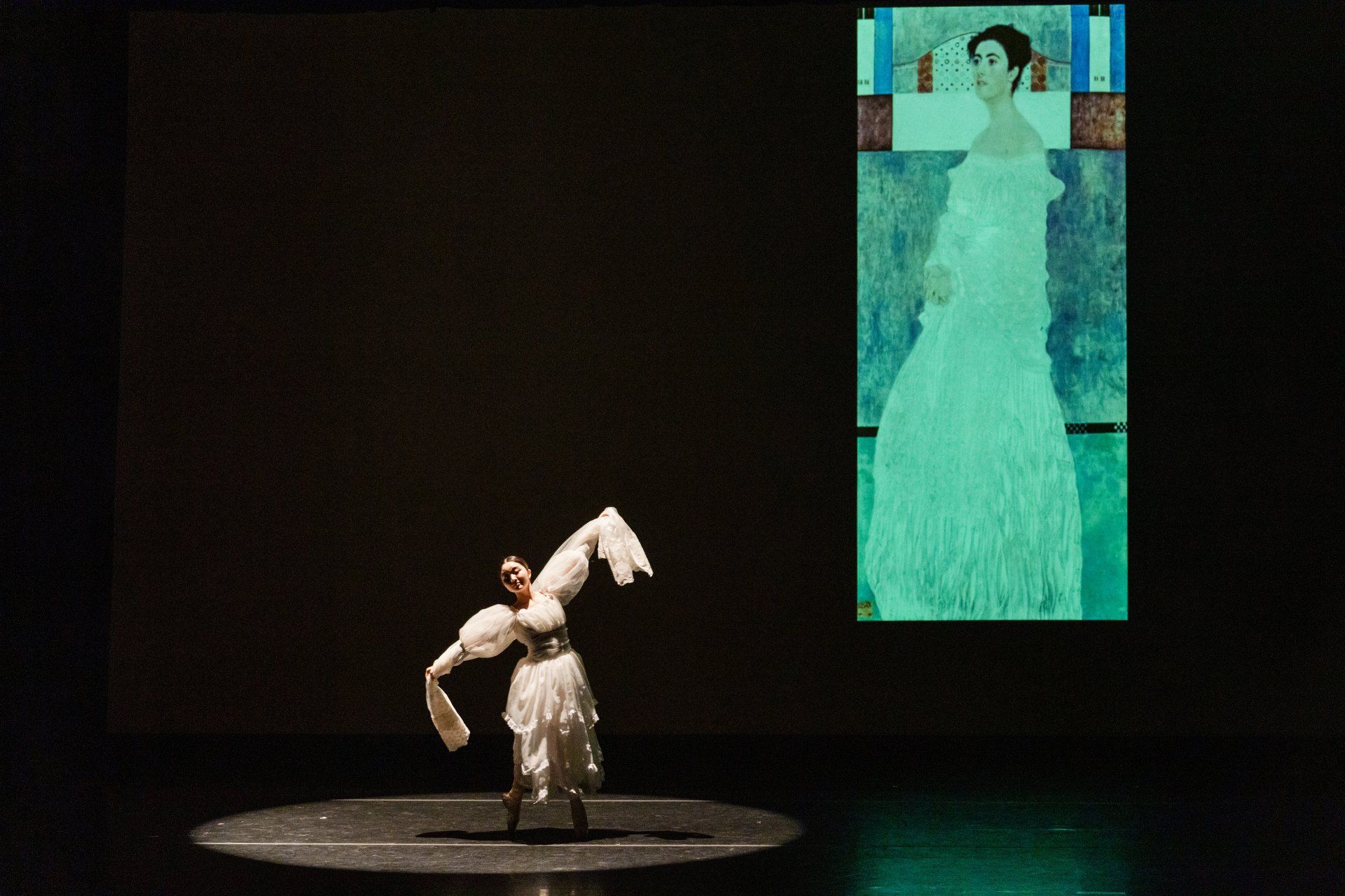 Performer in a silver dress stands onstage holding a white bird puppet, with a tall turquoise portrait projected behind.