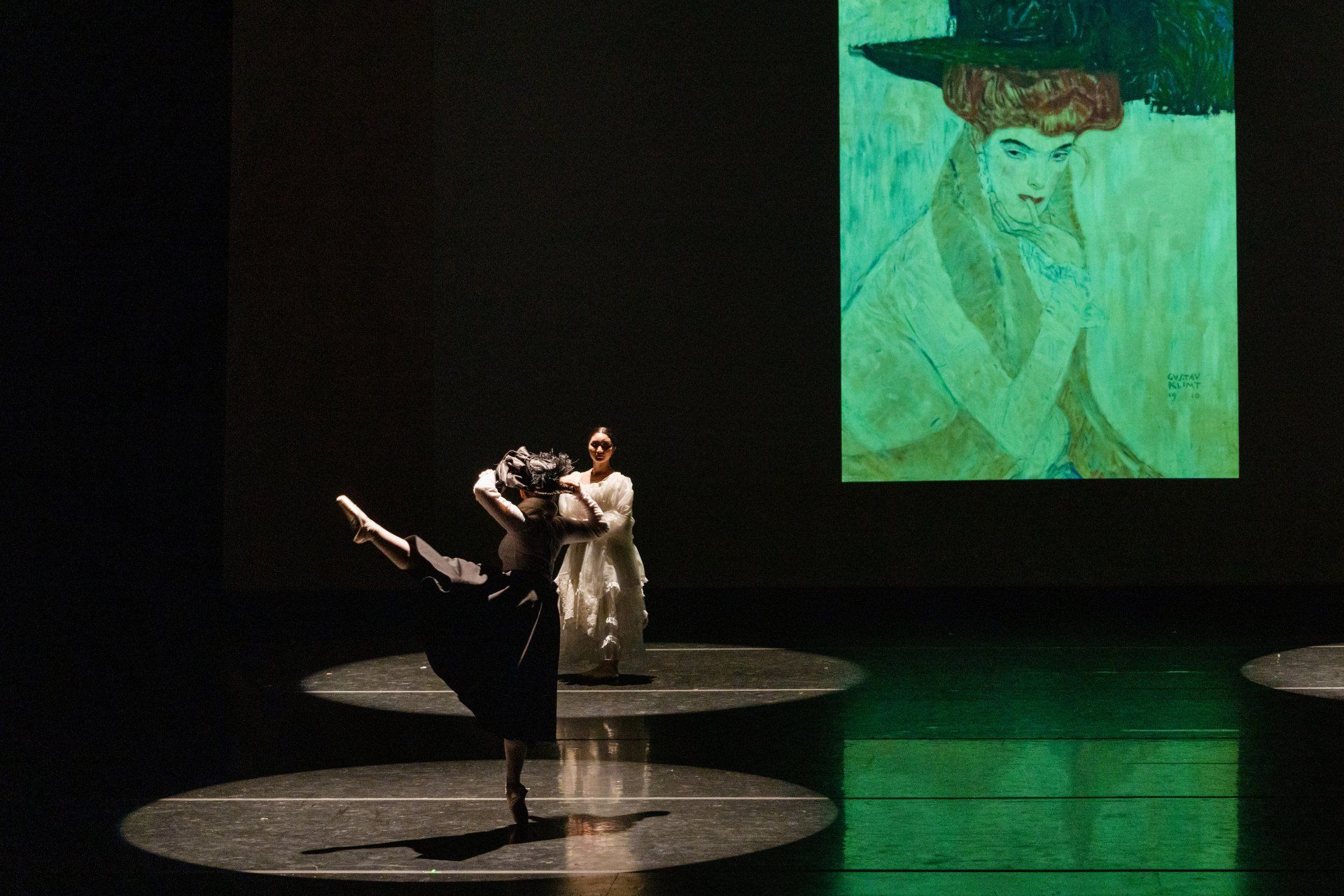 Two dancers in black on a dark stage, one lifting the other, with a green portrait projected behind them