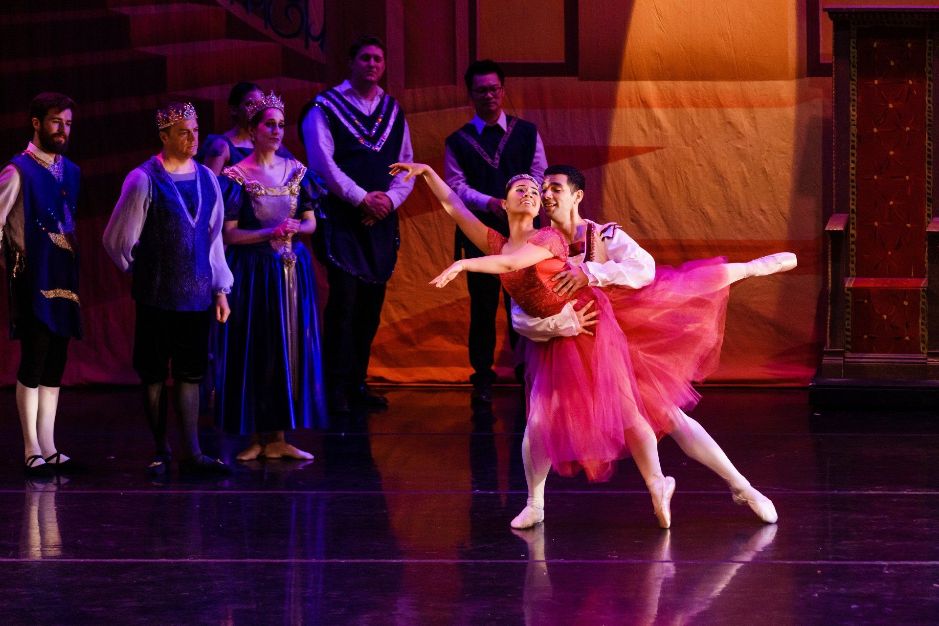 Two ballet dancers in pink costumes performing onstage, with other dancers watching in the background