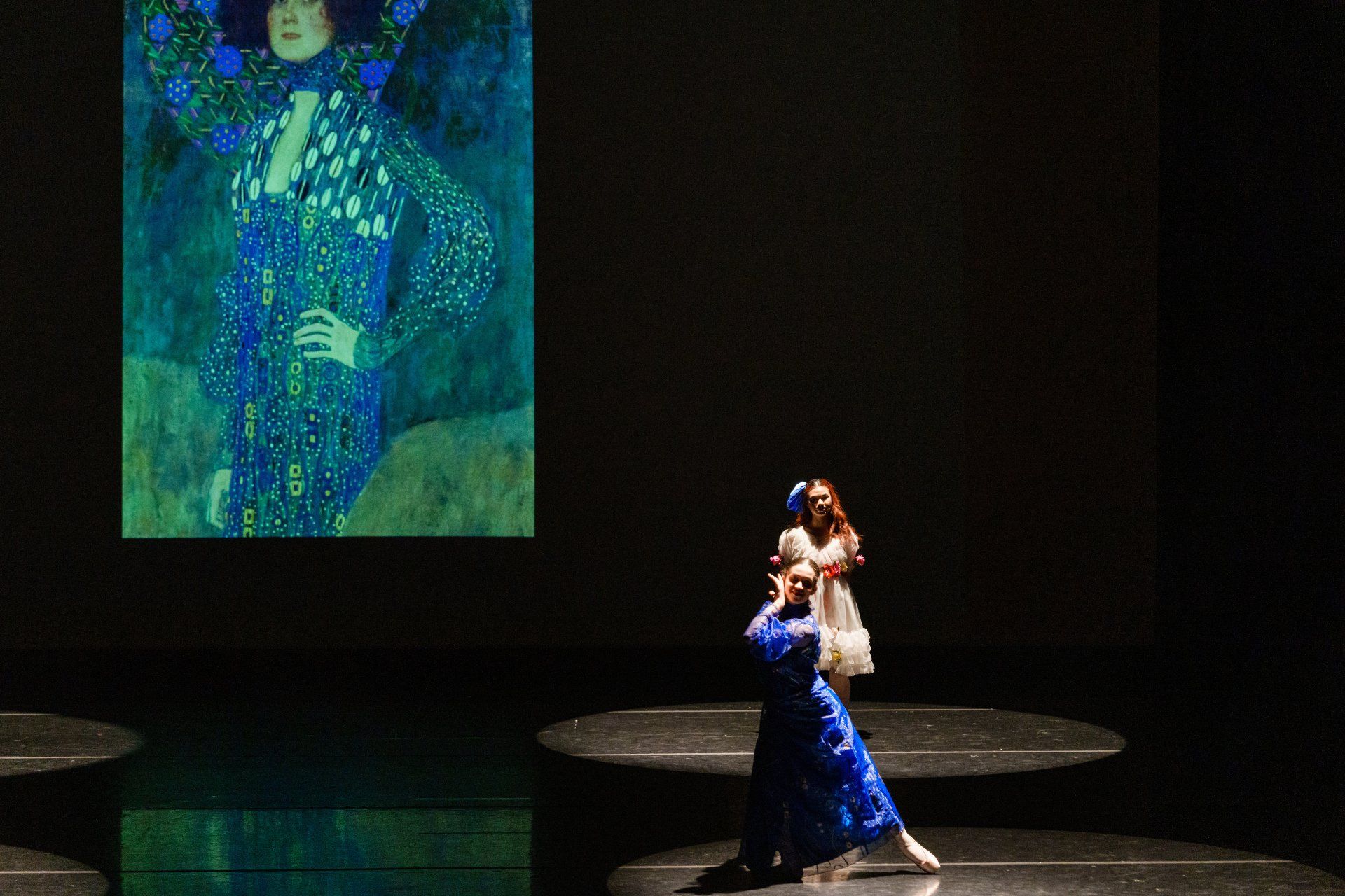 Dancer in a blue dress on a dark stage with a large projected portrait behind her