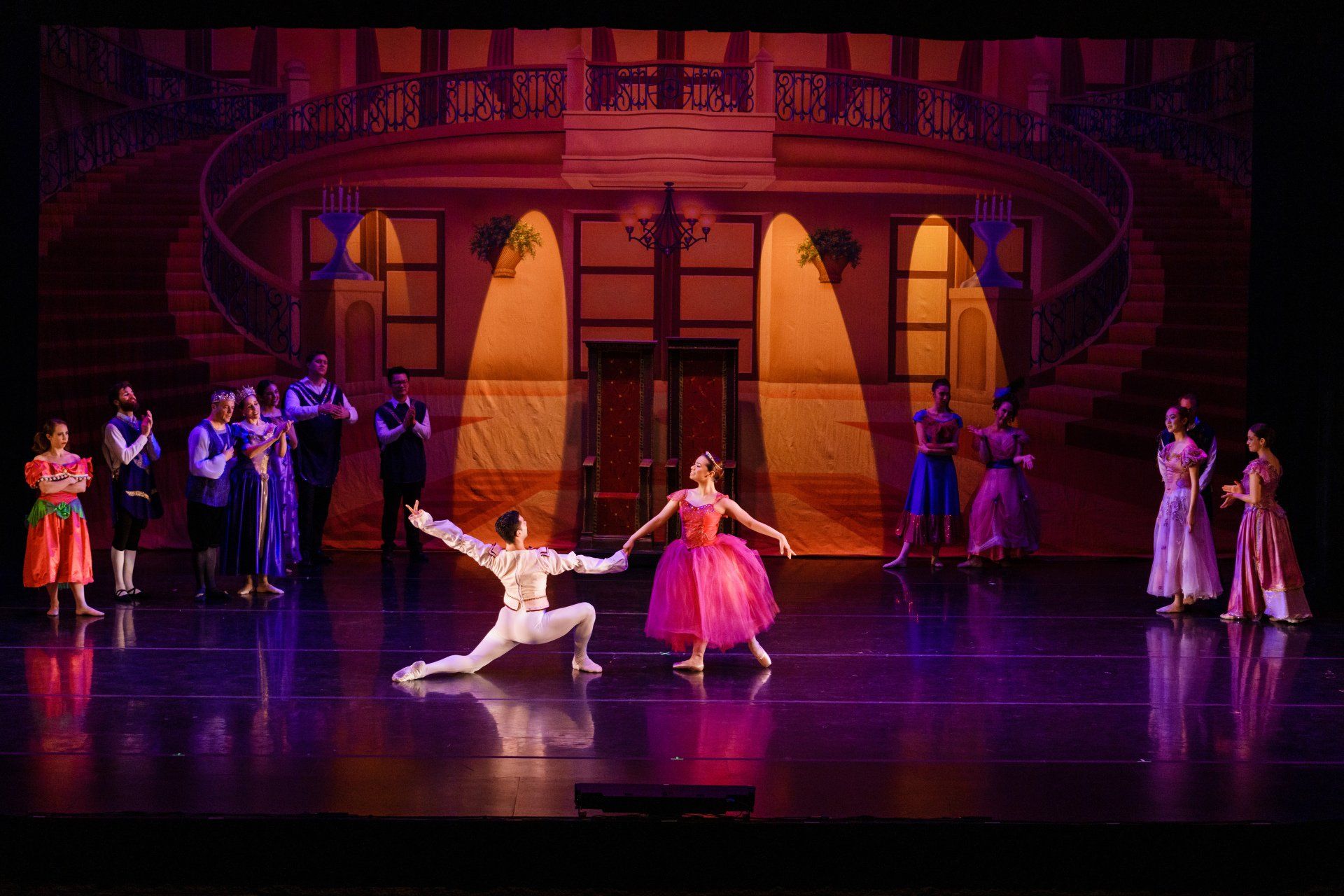 Ballet dancers on a stage with a pink-lit ballroom backdrop; one dancer in white and one in pink center stage.