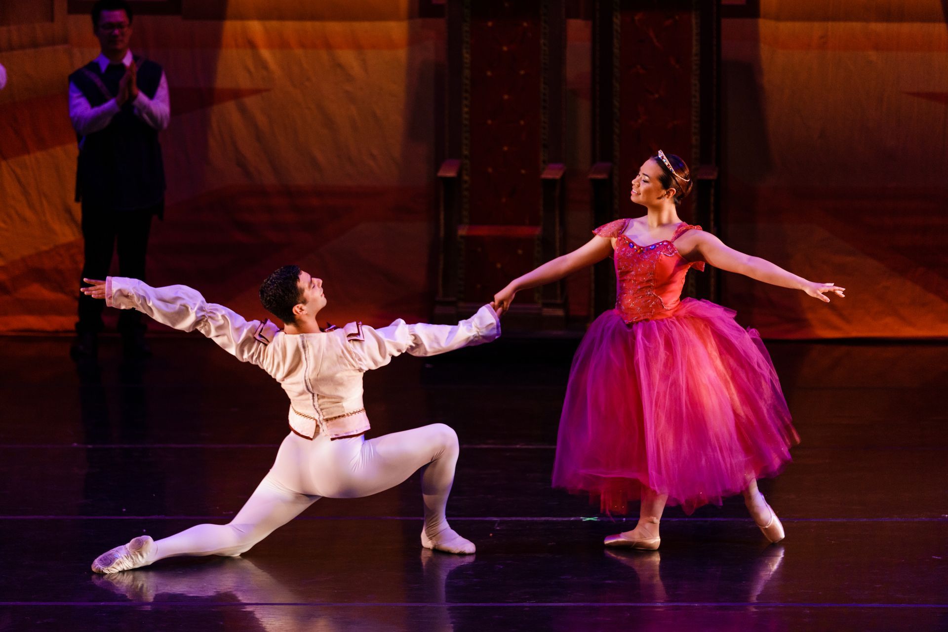 Two ballet dancers perform on stage, one kneeling in white and one standing in a pink tutu, holding hands.