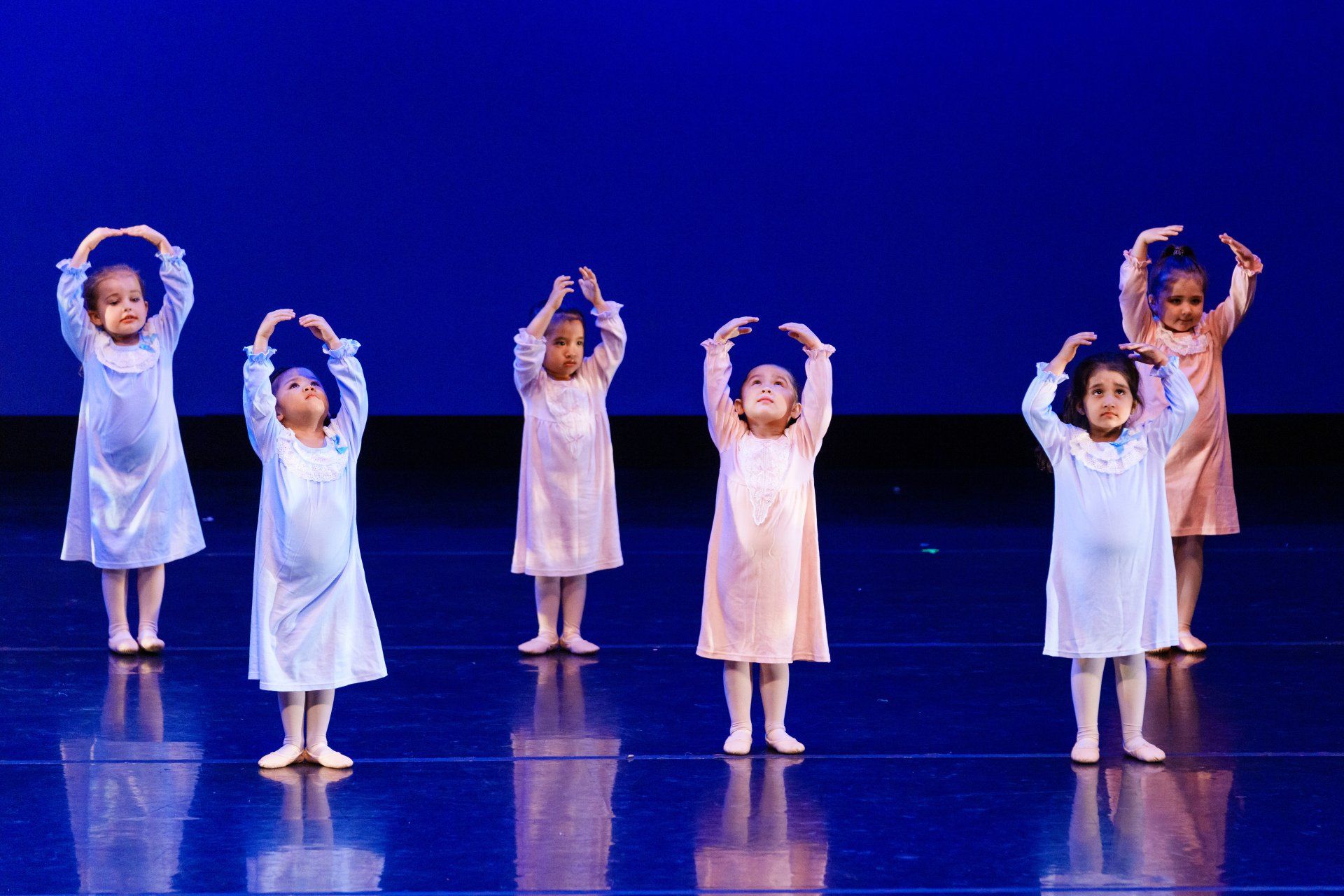Children in white dresses standing on a blue-lit stage with arms raised overhead