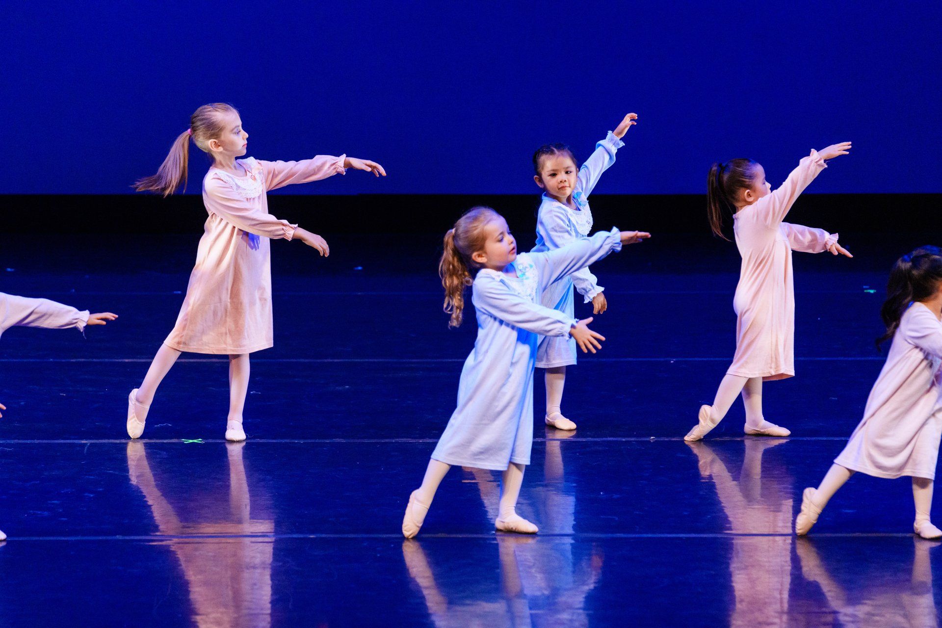Young dancers in pale costumes performing on a blue-lit stage with arms extended