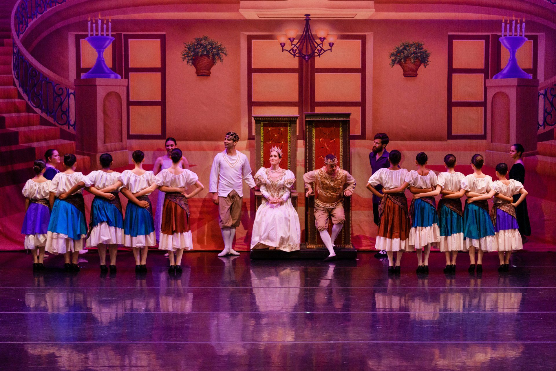 Stage performers in colorful costumes pose in formation in a theater set with purple lighting