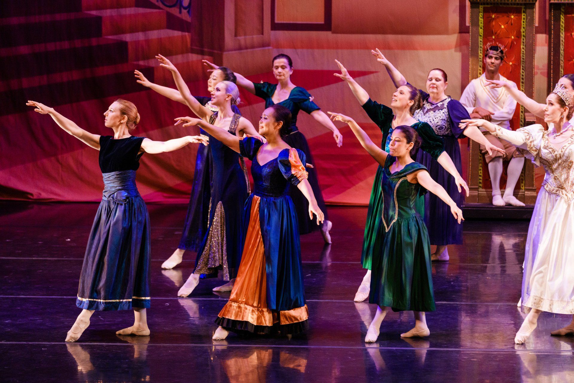 Costumed dancers on a theater stage, arms raised in a synchronized performance under pink lighting