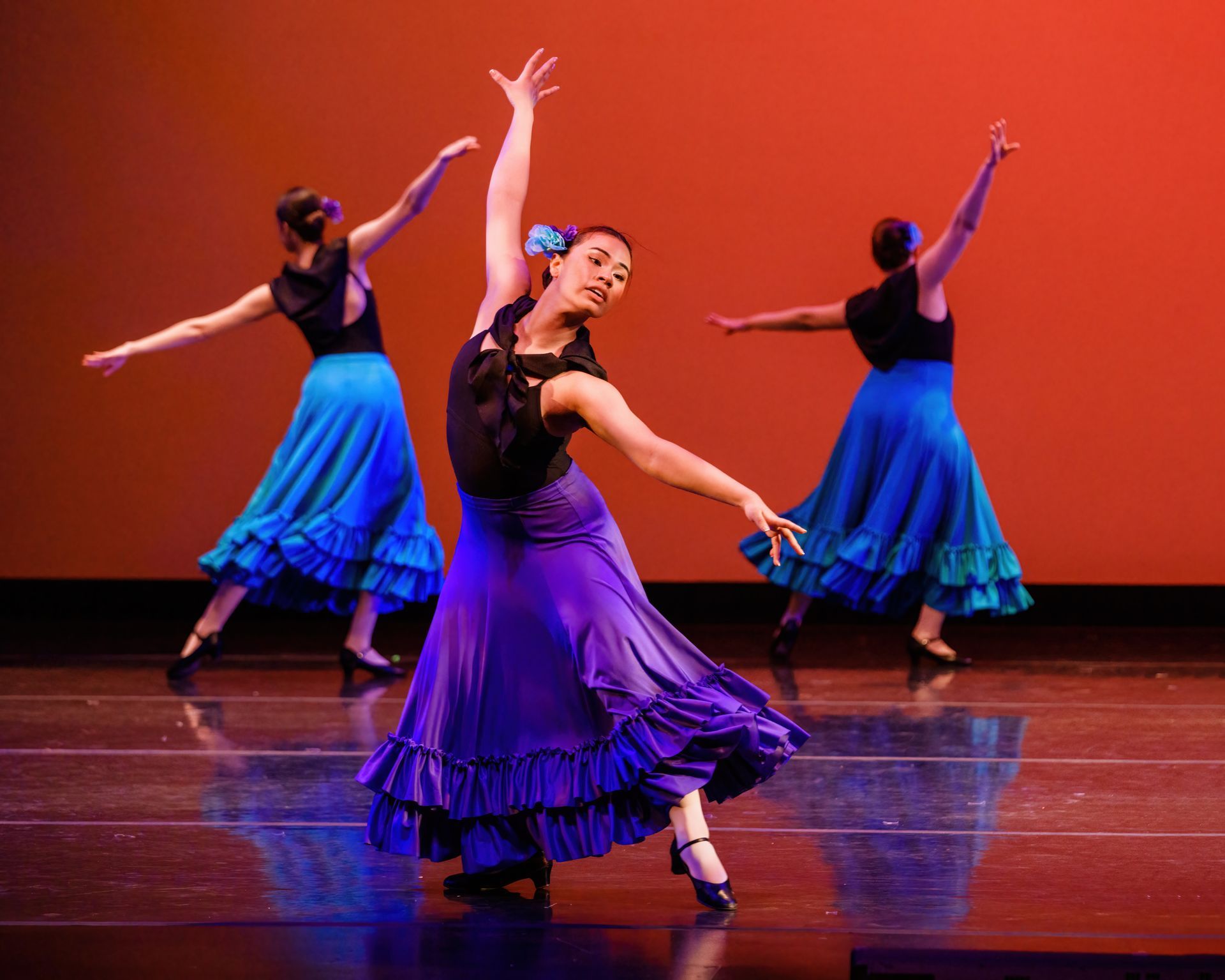 Three dancers in blue and purple dresses perform on a stage with a warm red backdrop.