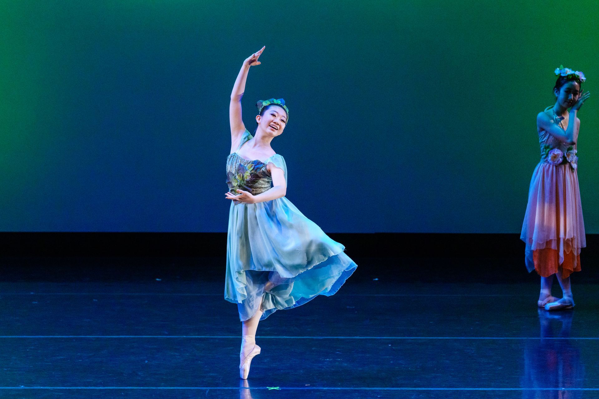 Ballet dancer in a blue costume performs on stage under colorful lighting, with another dancer in the background