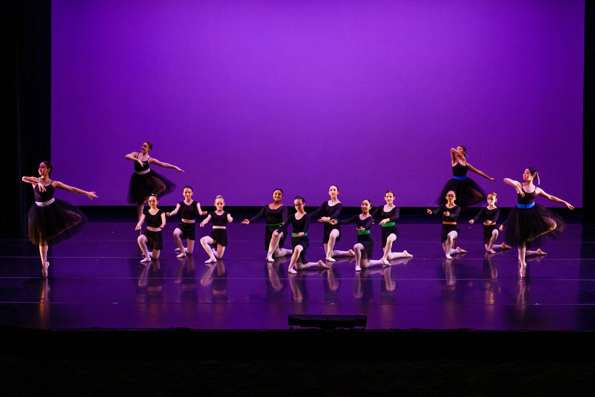 Dancers in black costumes perform on a purple-lit stage, with one dancer raised on each side.