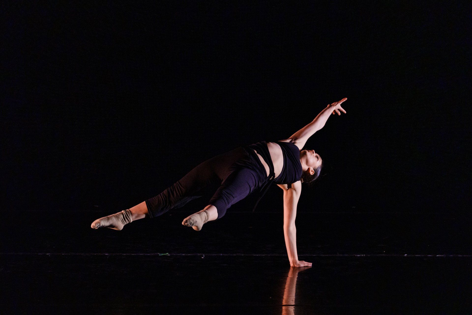 Dancer in a side plank pose on a dark stage, wearing dark athletic clothing with pink accents.