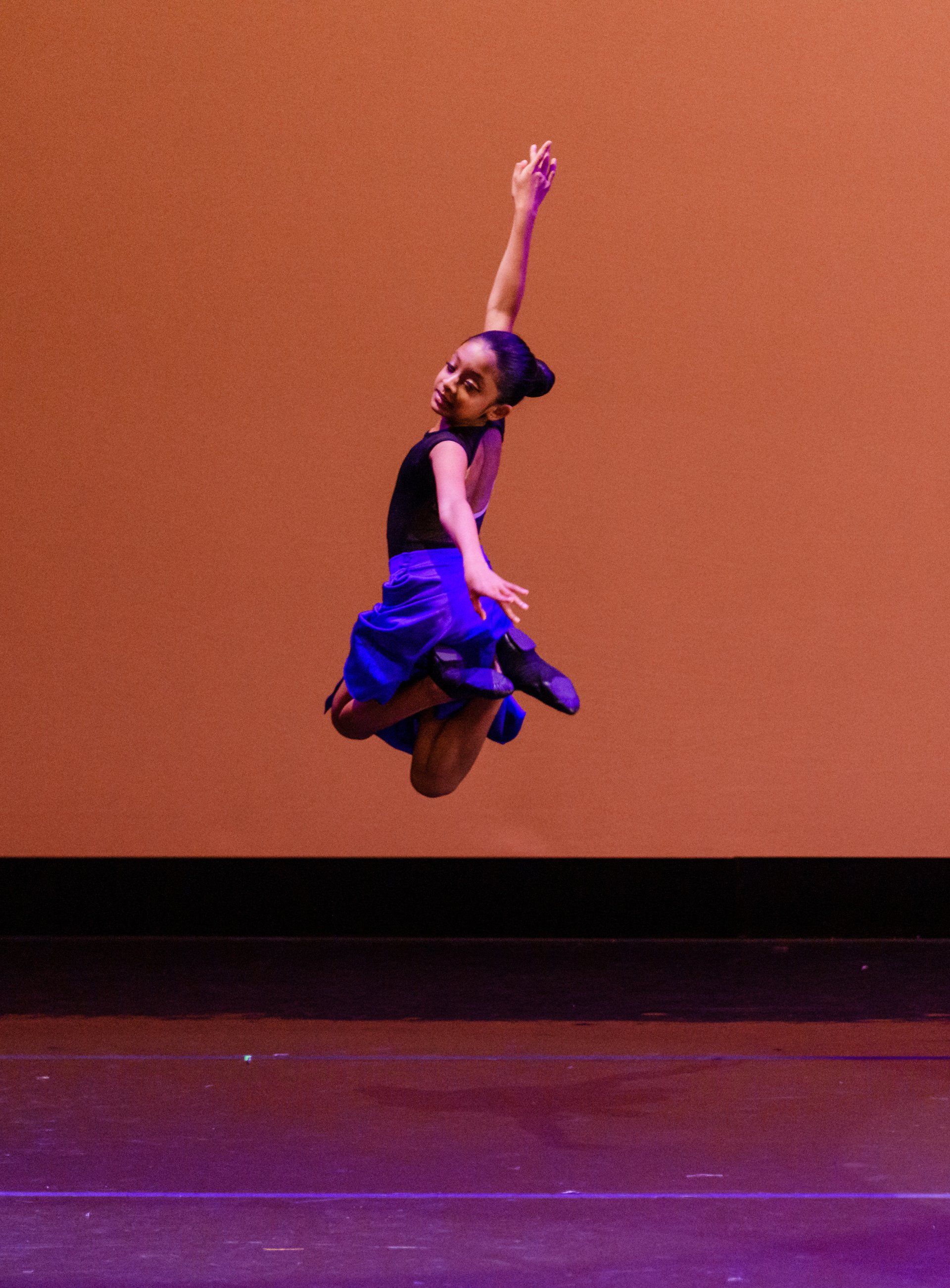 Dancer mid-air leap against an orange backdrop, wearing a black top and blue skirt.