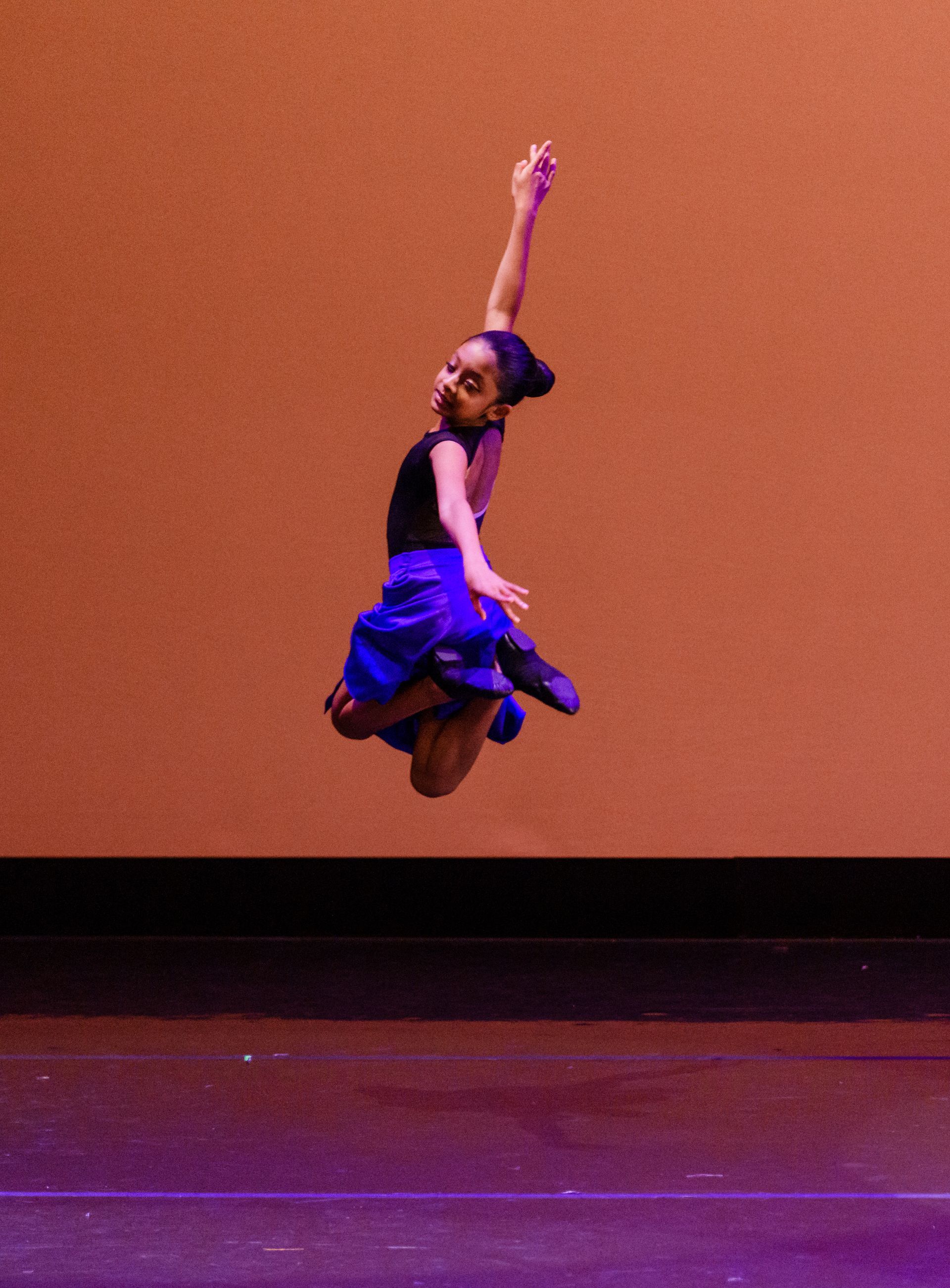 Dancer mid-air on a stage, arm raised, wearing a black top and blue skirt against an orange backdrop