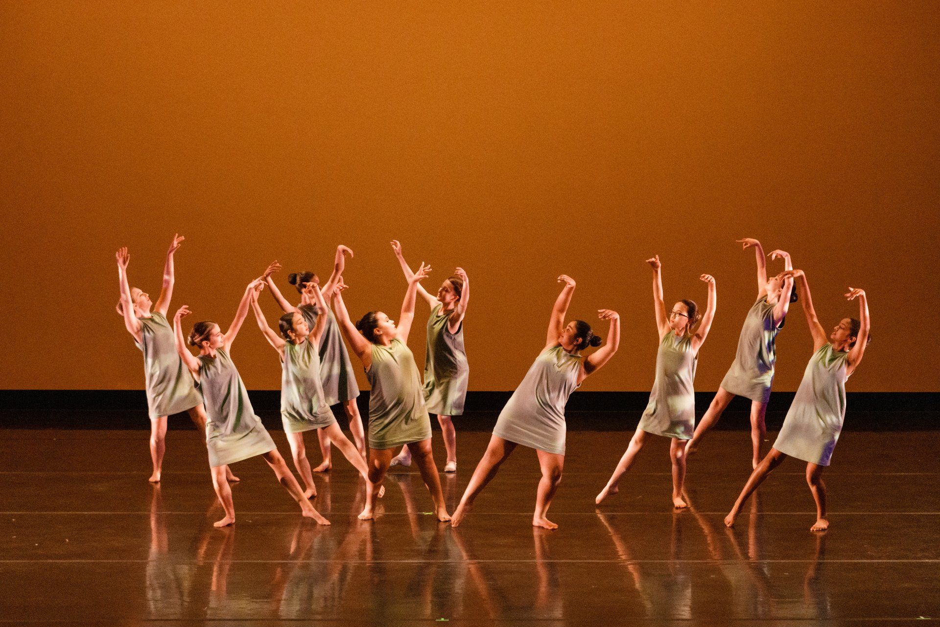 Dancers in gray dresses performing a synchronized routine on a stage with an orange-lit backdrop.