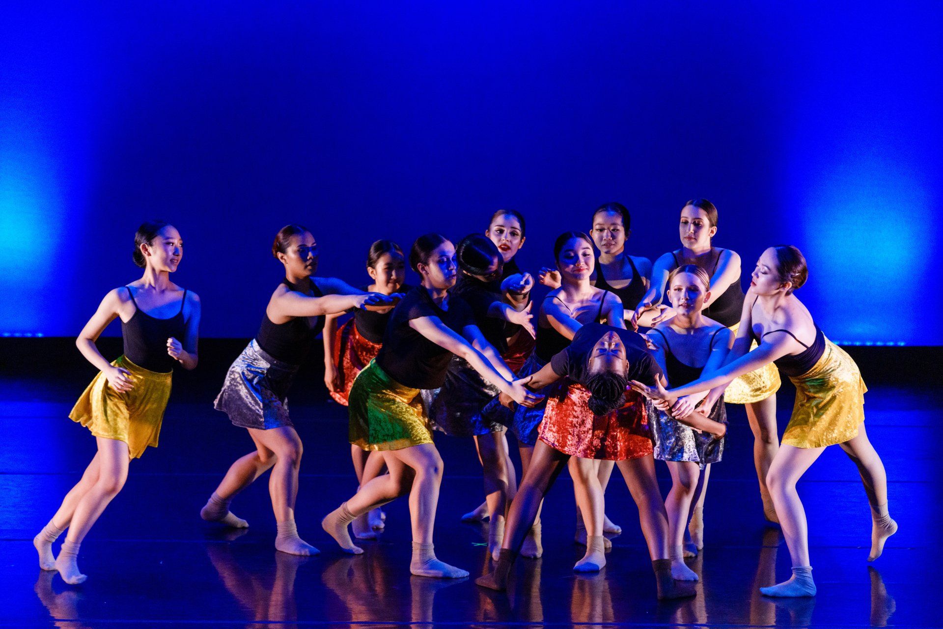Group of dancers performing on a blue-lit stage, wearing colorful costumes in synchronized poses