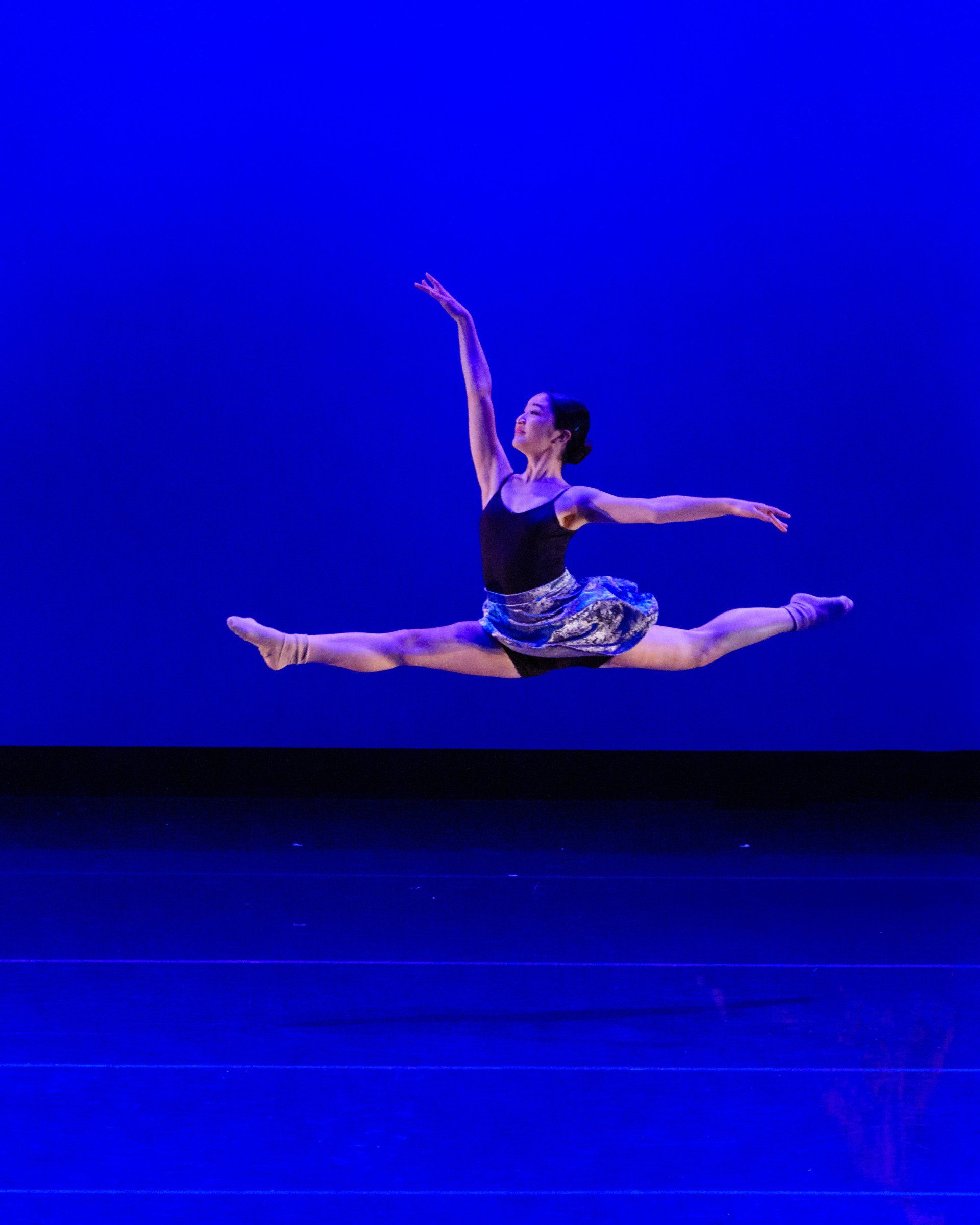 Dancer in a blue-lit stage leap, legs extended in a split and one arm raised.
