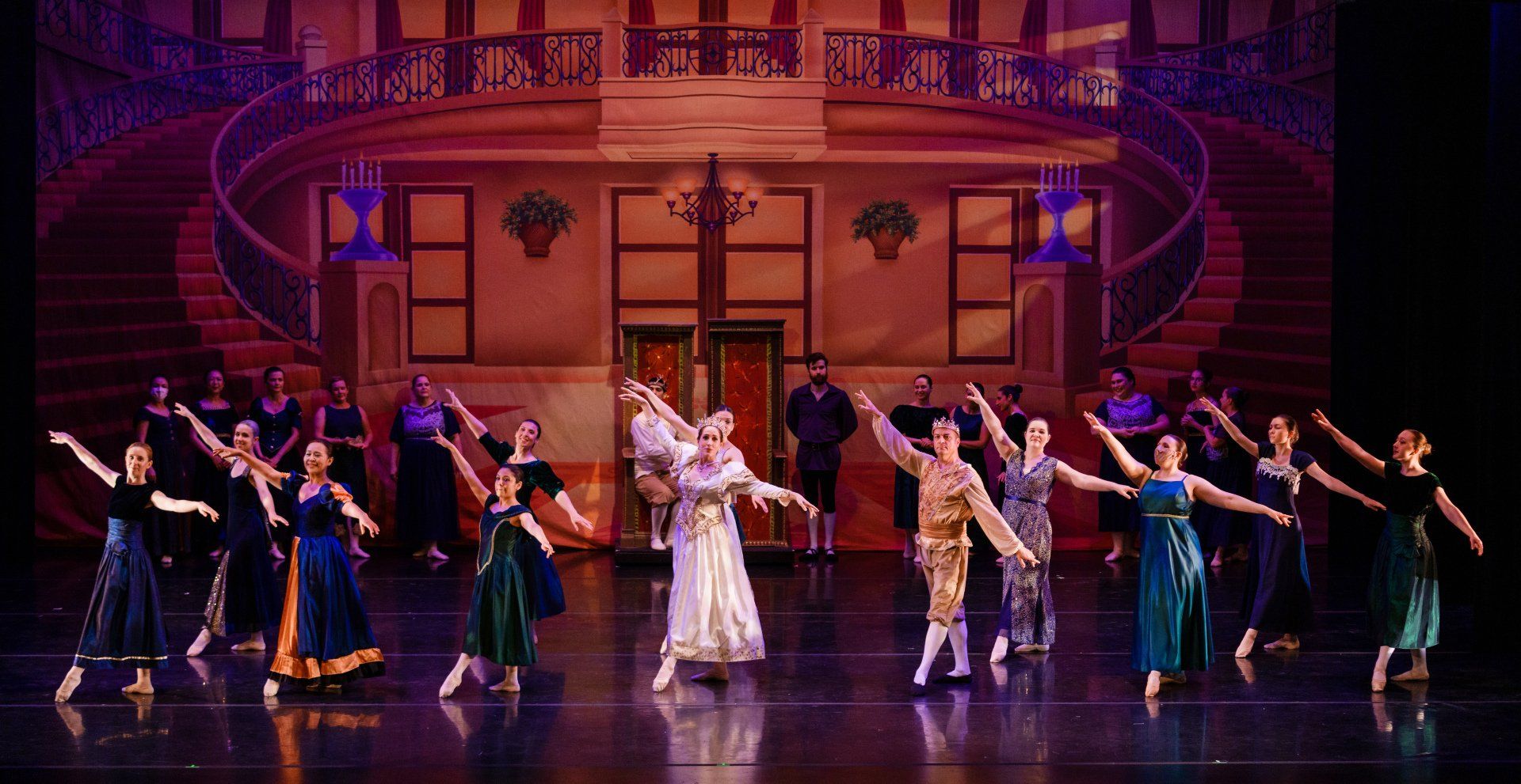 A line of dancers in colorful costumes on a stage with a grand staircase and warm purple lighting.