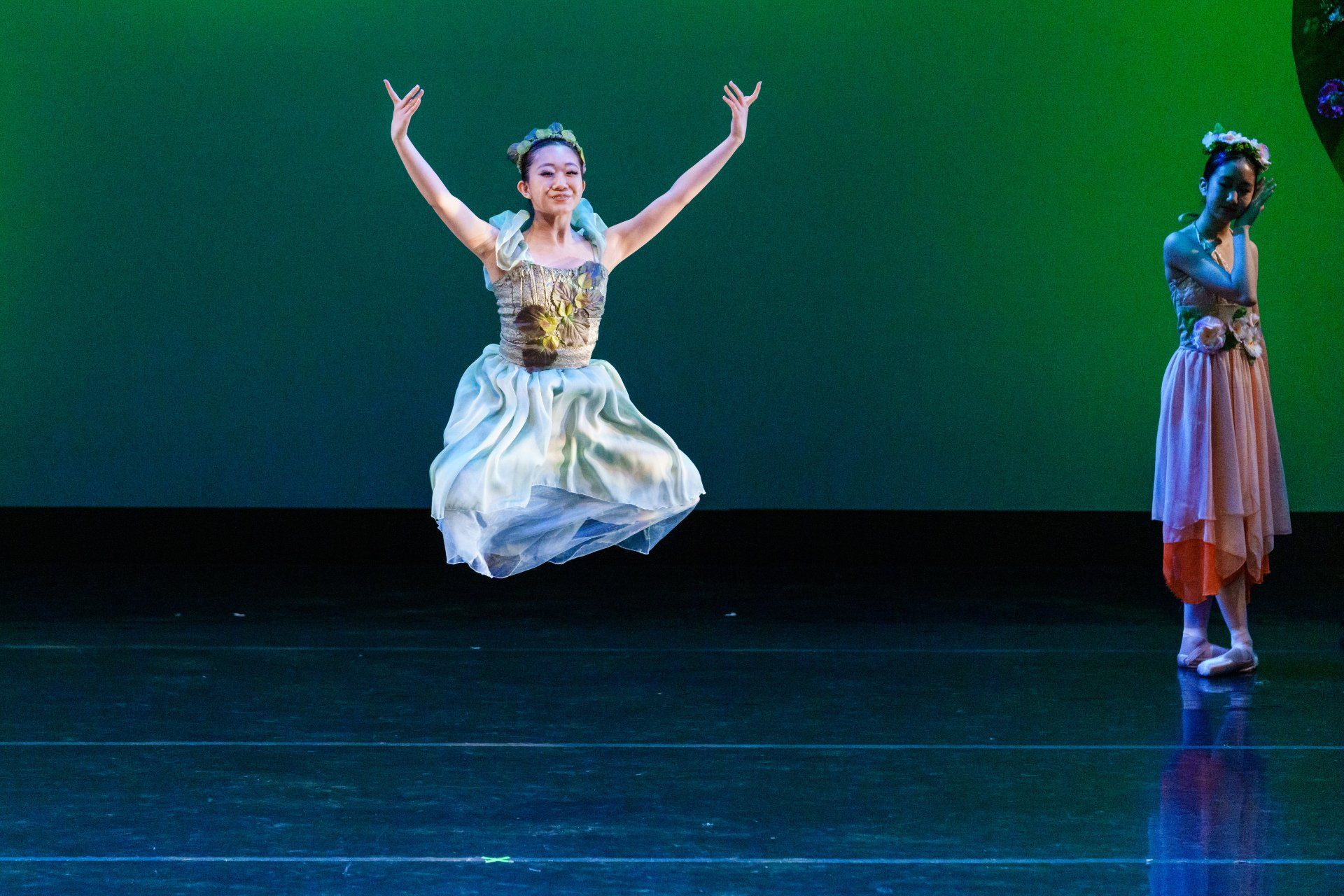 Two dancers on a stage with a blue-green backdrop, one mid-jump in a white dress