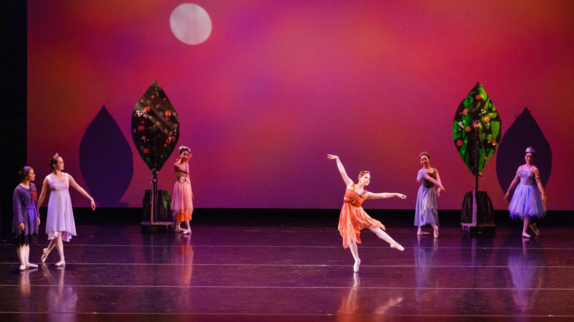 Ballet dancers perform onstage before a pink and purple backdrop with tall leaf-shaped props.