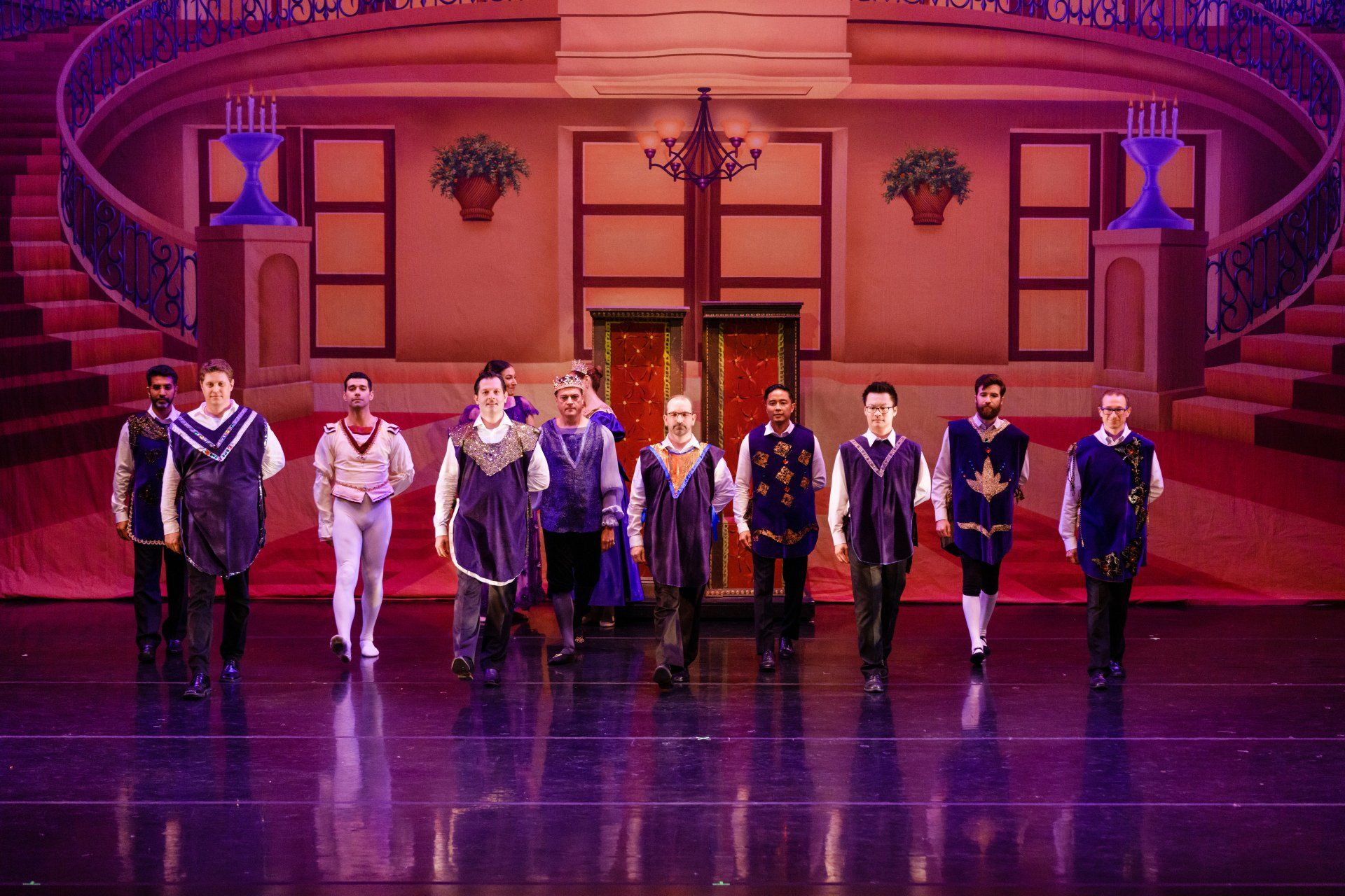 Nine performers stand on a purple-lit theater stage before a grand staircase backdrop.