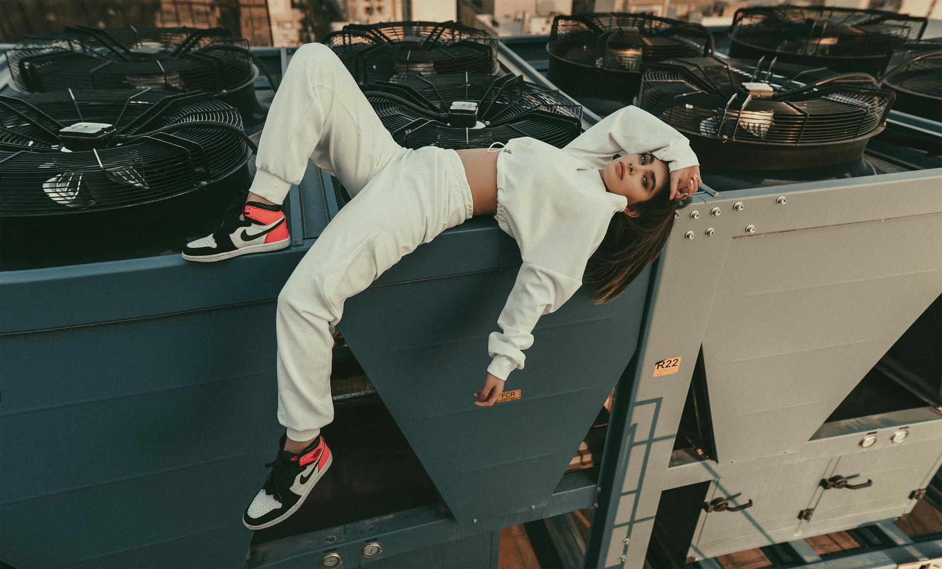 Woman in white sweatsuit and Nike sneakers lying on a rooftop HVAC unit, blending fashion with industrial elements.