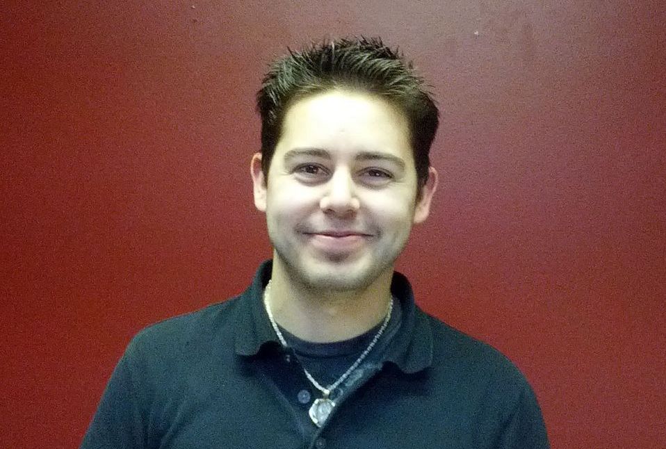 A man wearing a black shirt and a necklace is smiling in front of a red wall.