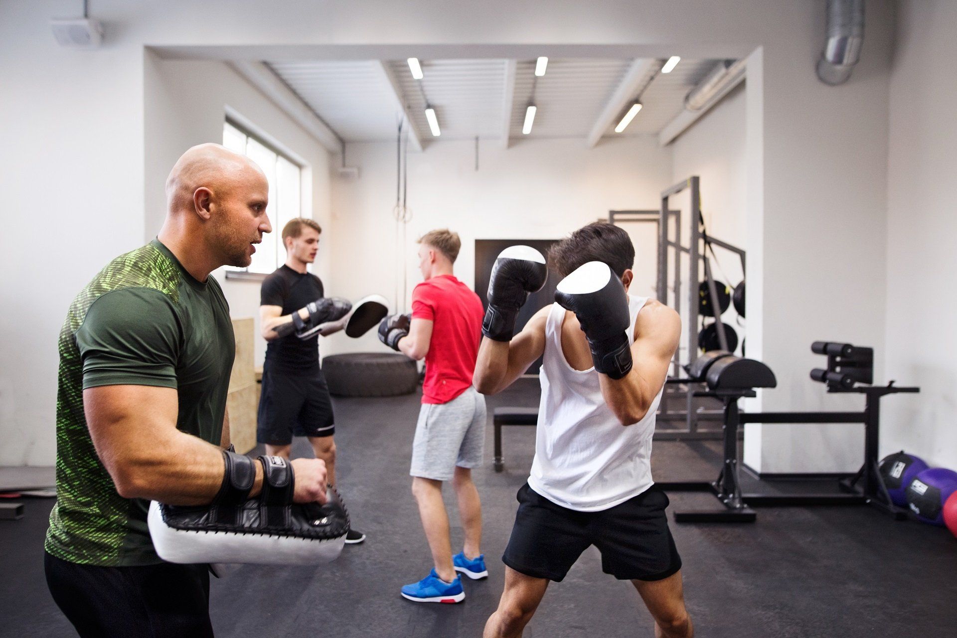 People boxing in a gym with a trainer holding mitts and others exercising in the background