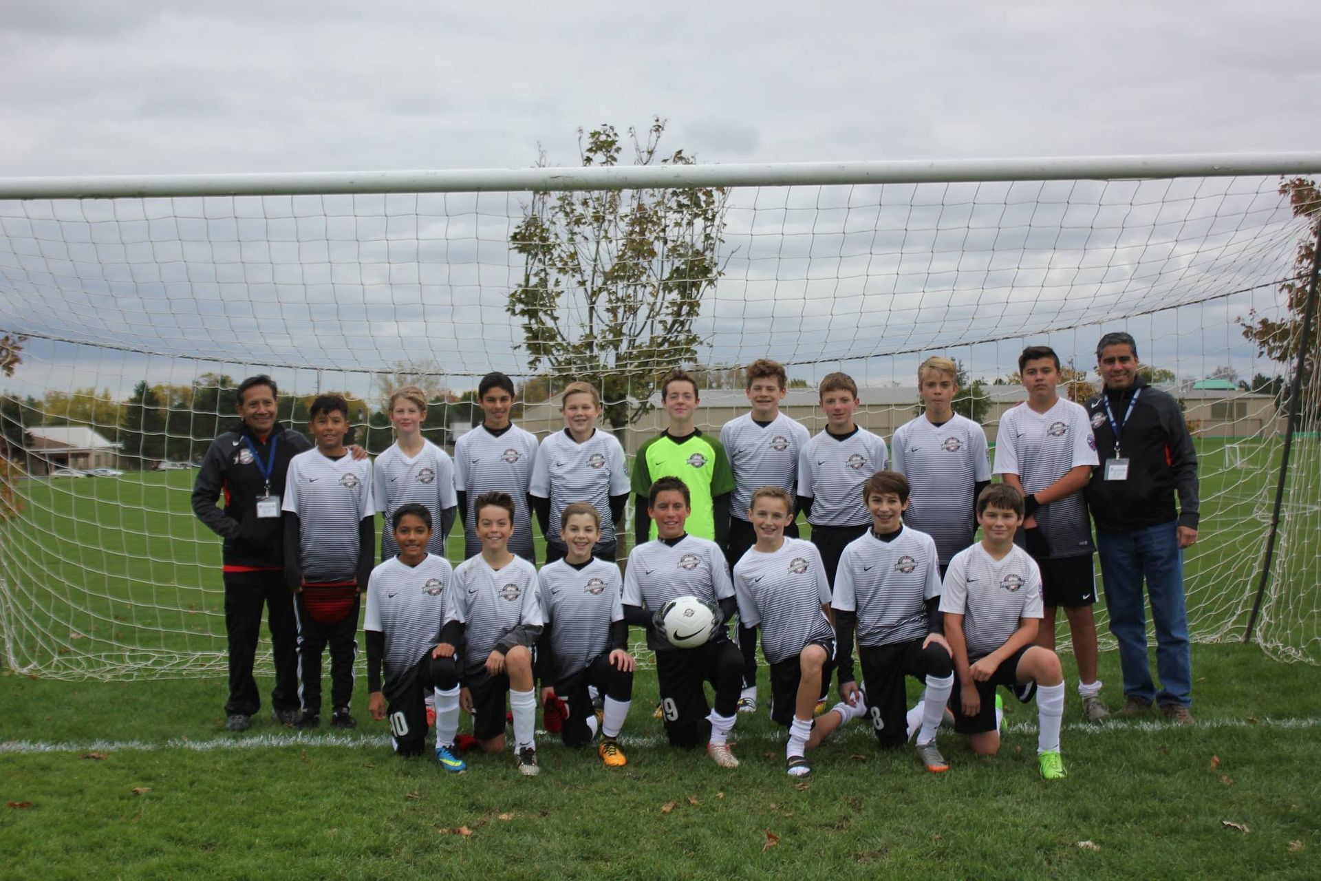 Soccer team posing in front of a goal on a grassy field, with coaches at the sides and a ball in front.