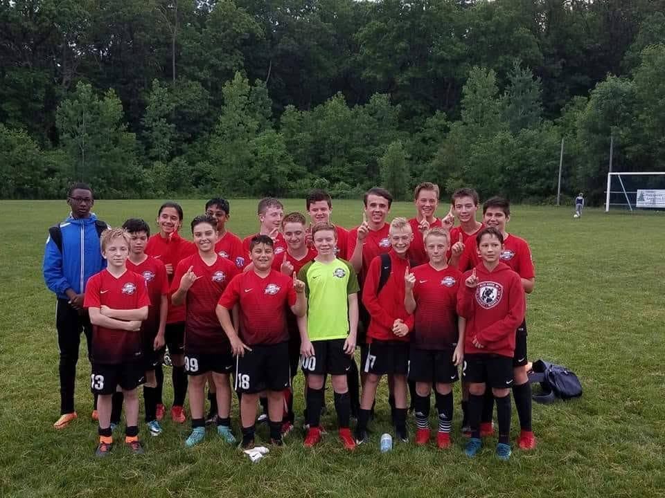 Youth soccer team in red jerseys posing on a grassy field with a coach and goal in the background