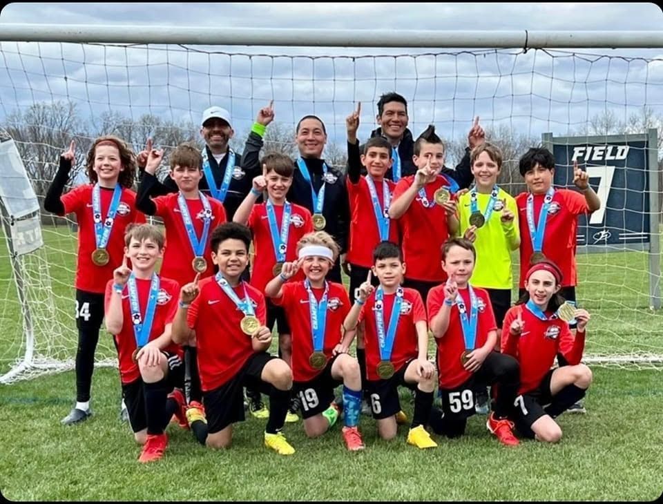 Youth soccer team in red jerseys posing with medals on a field, with coaches behind them.