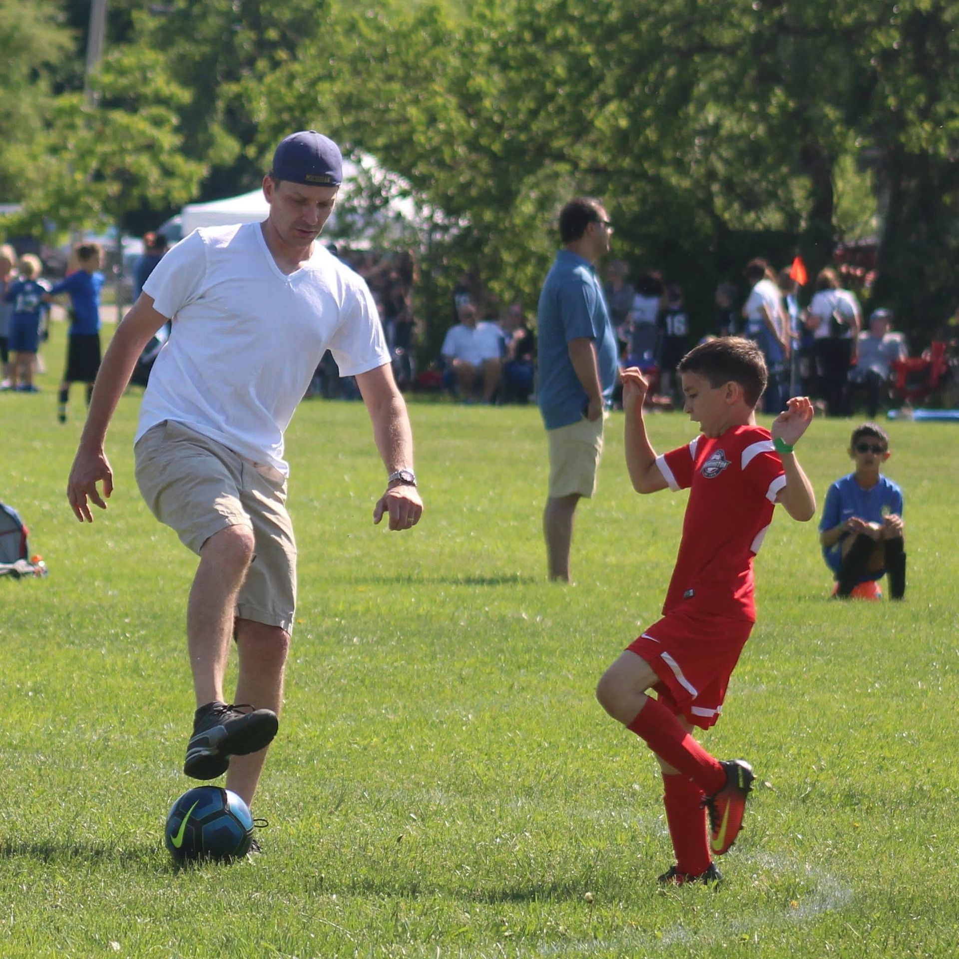 Adult and child playing soccer on a sunny grassy field with spectators in the background
