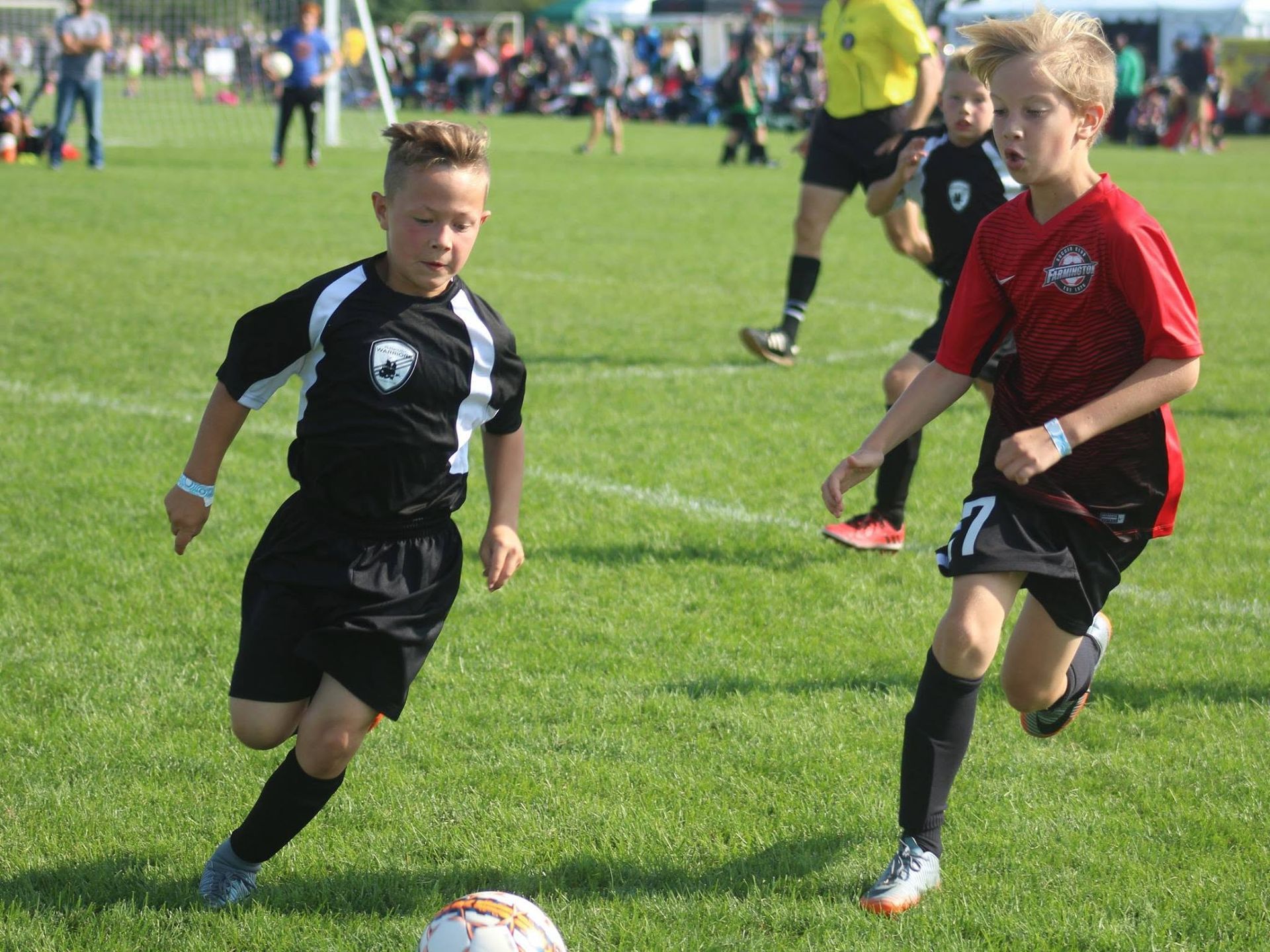 Two boys playing soccer on a grassy field, one in black and one in red, with a goal in the background