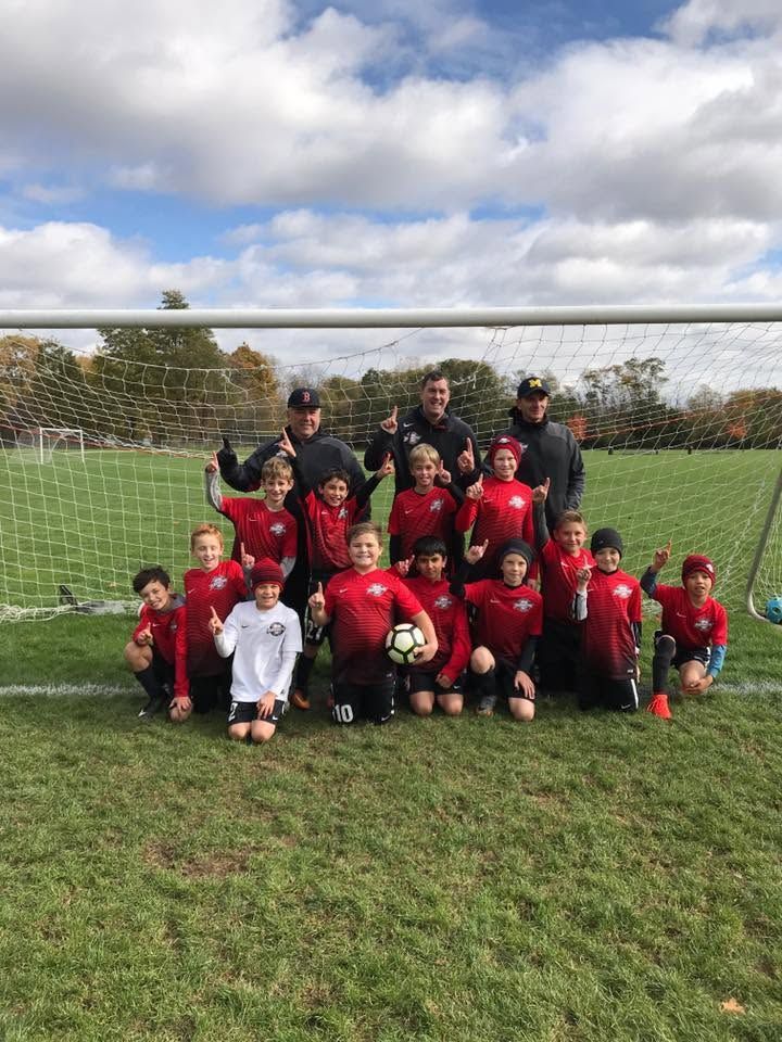 Youth soccer team posing on a grassy field in front of a goal, holding a ball and wearing red and black uniforms.