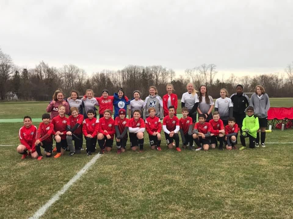 Youth soccer team posing on a field, with red uniforms in front and gray-clad players and coaches behind.