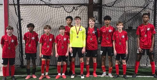 Youth soccer team in red jerseys posing on an indoor field, with a coach in a yellow shirt at center.