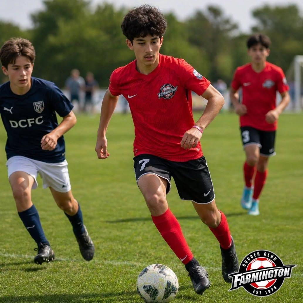 Soccer players in red and navy uniforms sprint toward the ball on a grassy field.