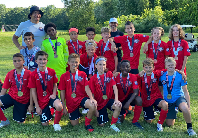 Youth soccer team posing on a grassy field with medals and trophies, wearing red jerseys after a match.