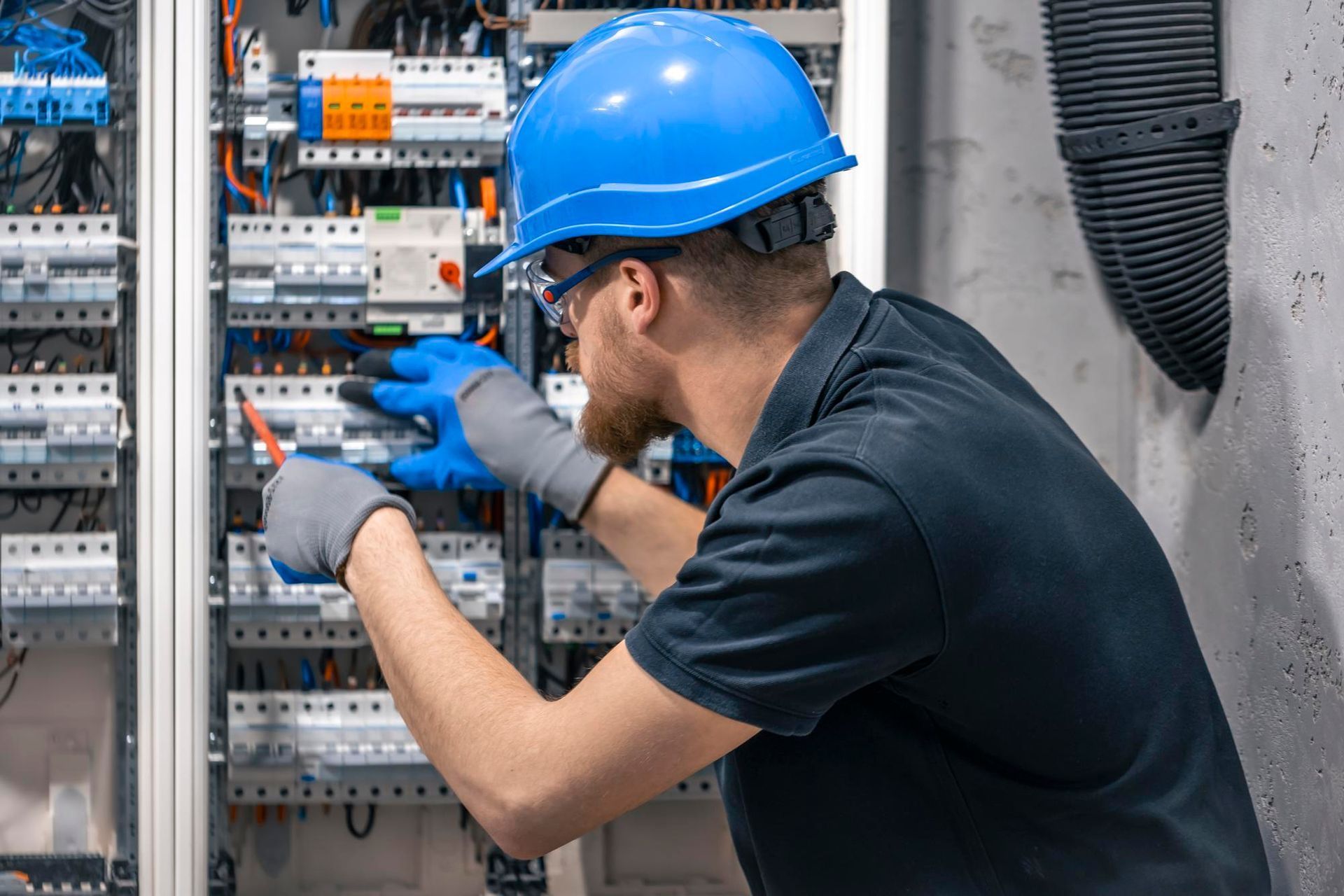 Electrician in blue hard hat, safety glasses, and gloves working on an electrical panel.