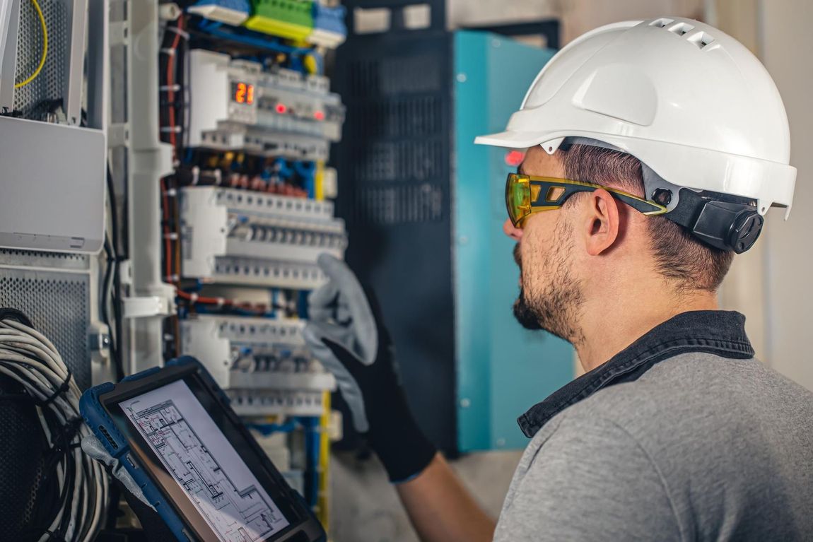Electrician inspecting electrical panel, wearing safety glasses and a hard hat, looking at a tablet.